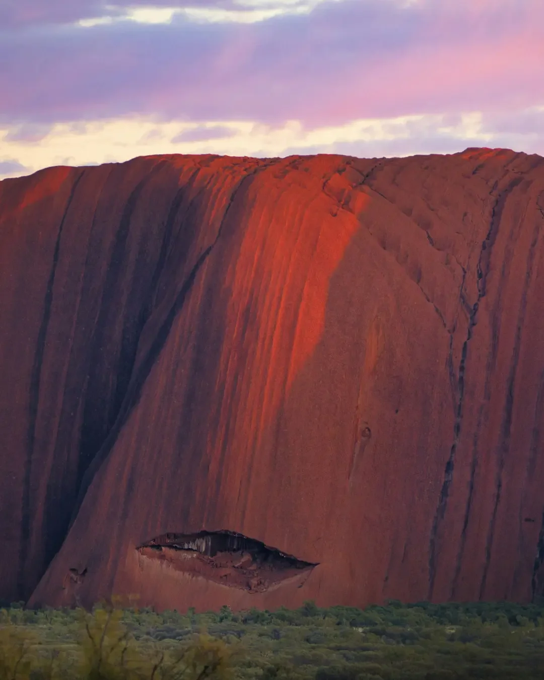 A large, reddish-brown rock formation with a natural arch formation near its base. The sky above has a purple and pink hue with some clouds, and there is green foliage at the bottom of the image.