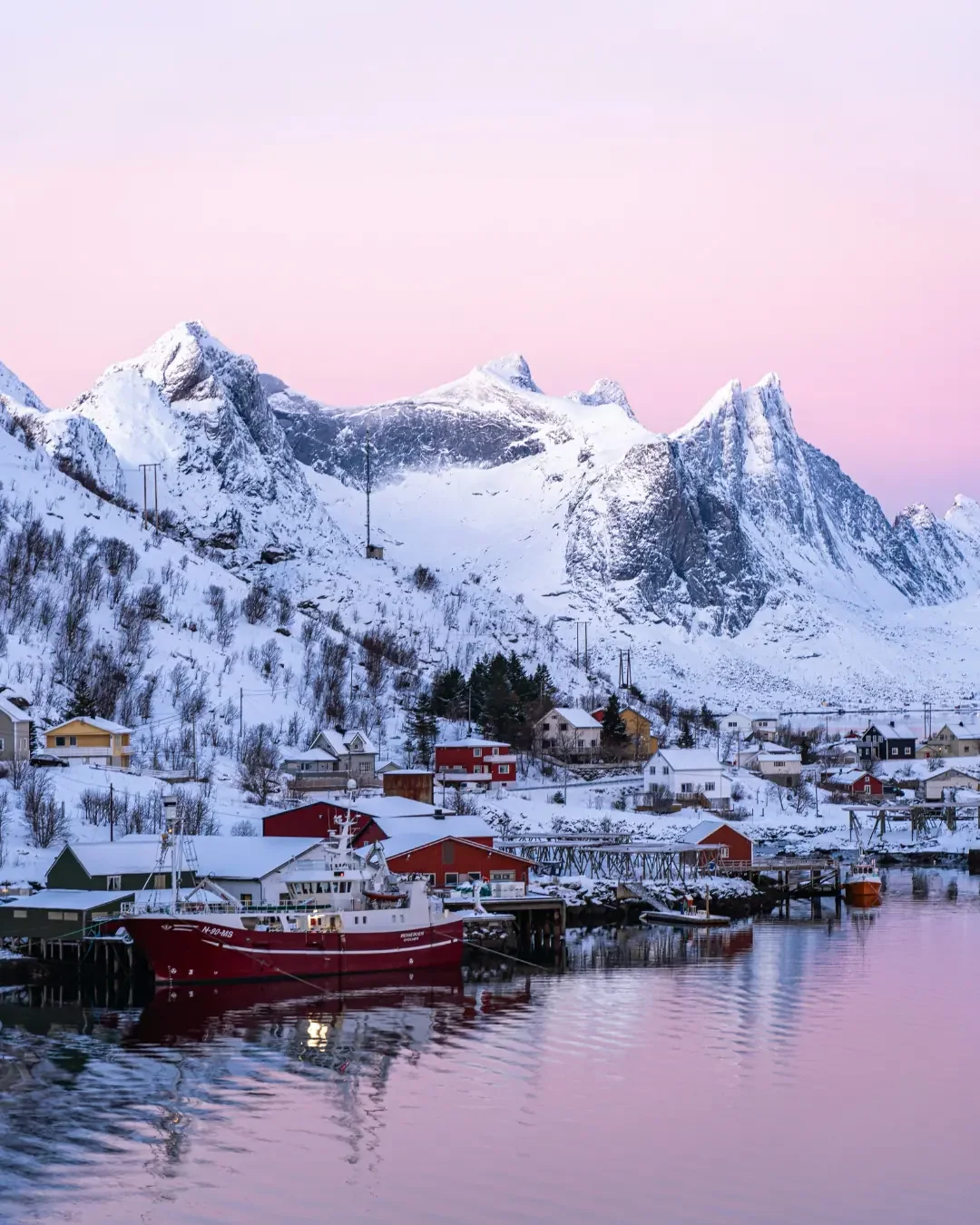 A picturesque view of Hamnøy fishing village in the Lofoten Islands at sunset, featuring traditional red rorbu cabins and the dramatic peak of Mount Olstinden in Norway.