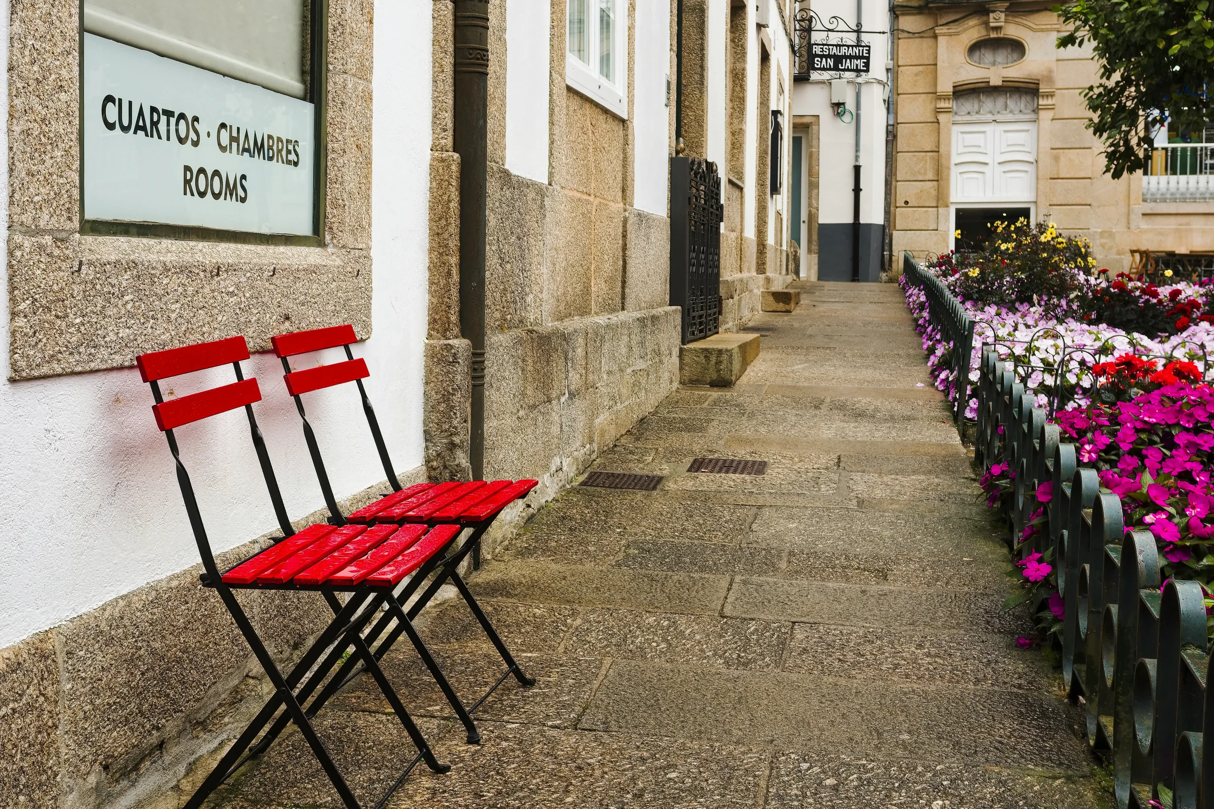 Exterior view of a traditional stone pilgrim albergue with a wooden door, a yellow arrow sign on the wall, and backpacks resting near the entrance.