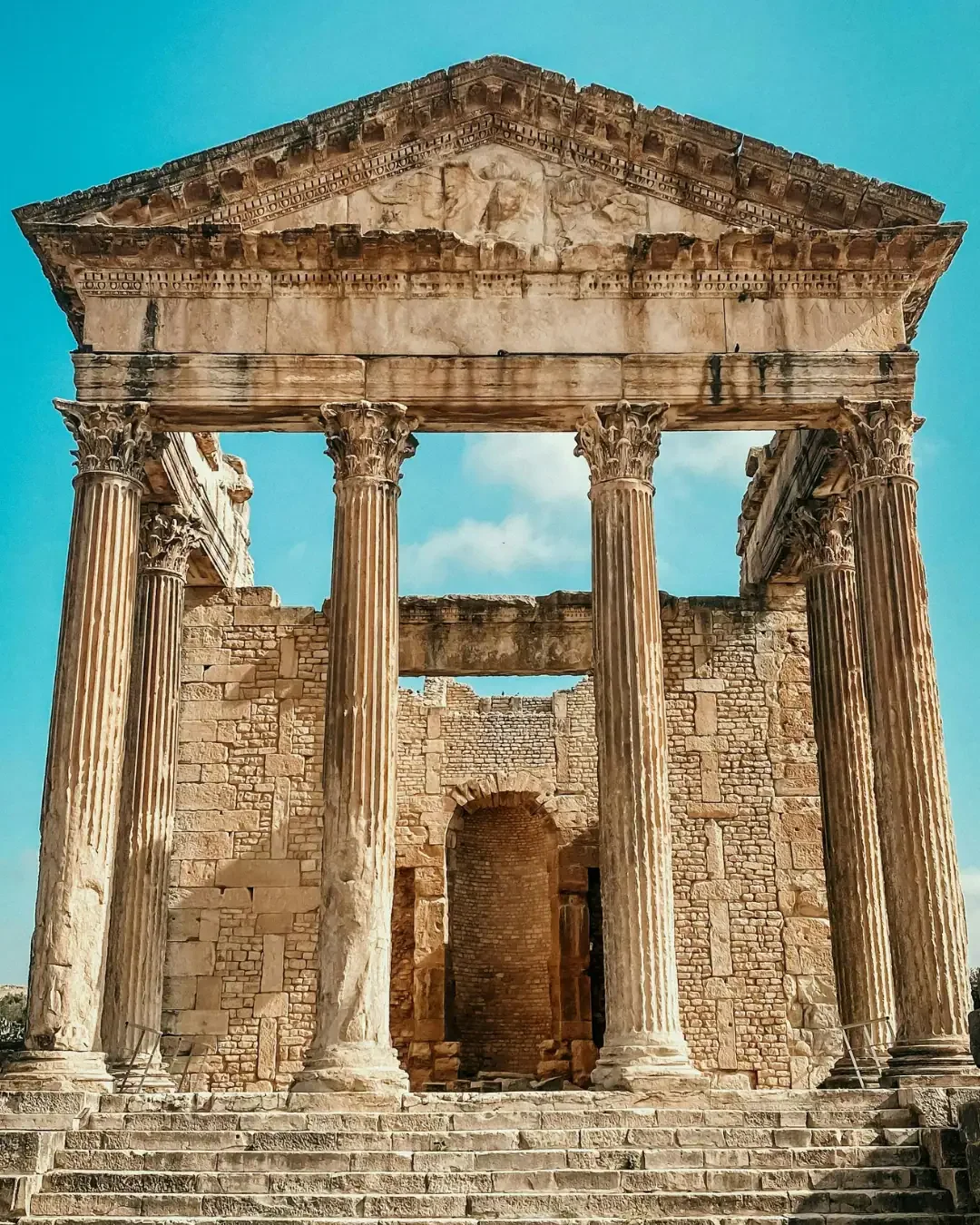 The well-preserved Capitol Temple at the Dougga archaeological site in Tunisia, featuring grand Corinthian columns and a triangular pediment under a bright blue sky.