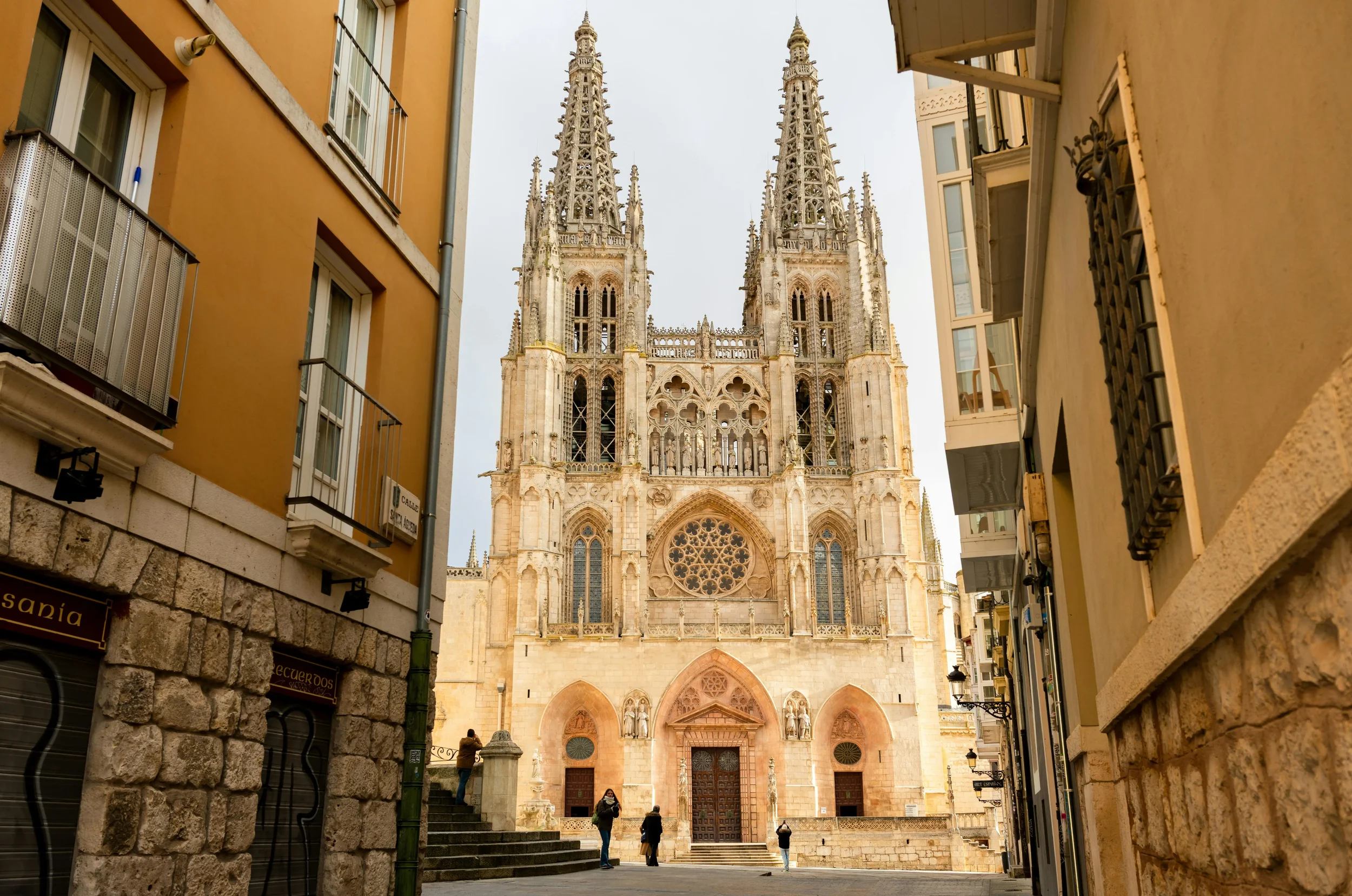 Impressive facade of the Gothic Burgos Cathedral in Spain with its twin openwork spires and ornate stone carvings against a blue sky.