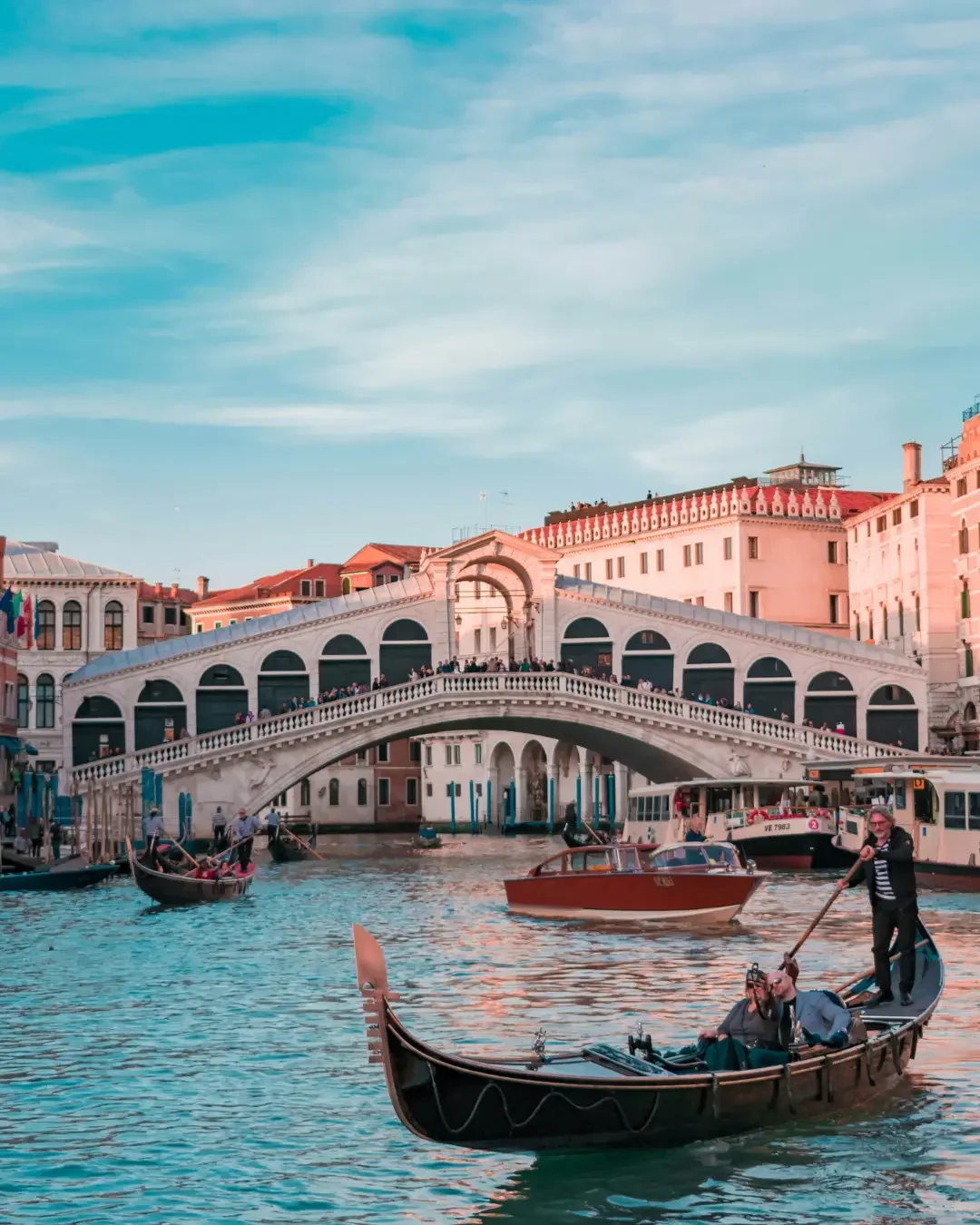 View of the Rialto Bridge over the Grand Canal in Venice, Italy, with boats and gondolas on the water under a partly cloudy sky.