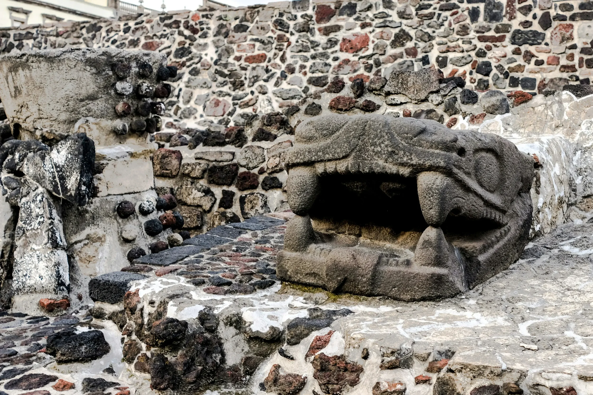 Stone ruins of the Templo Mayor archeological site with the Metropolitan Cathedral and colonial buildings in the background under a clear sky in Mexico City.