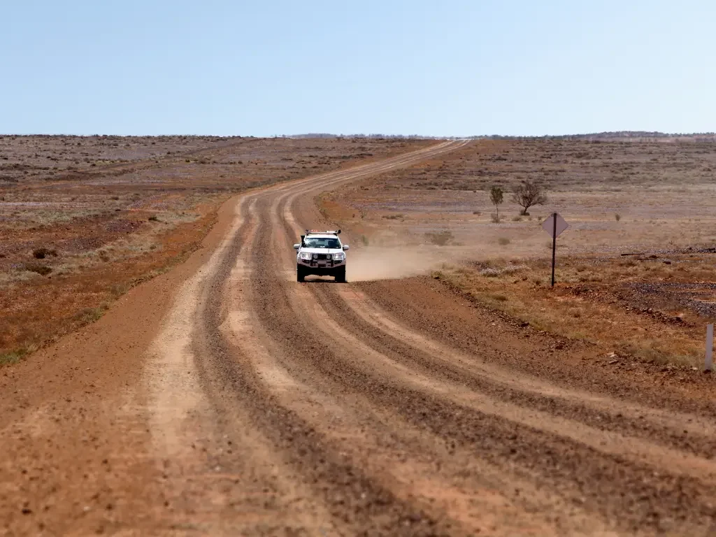 An off-road 4WD vehicle kicking up red dust while driving on a rugged dirt track through the vast and arid landscape of the Australian desert.