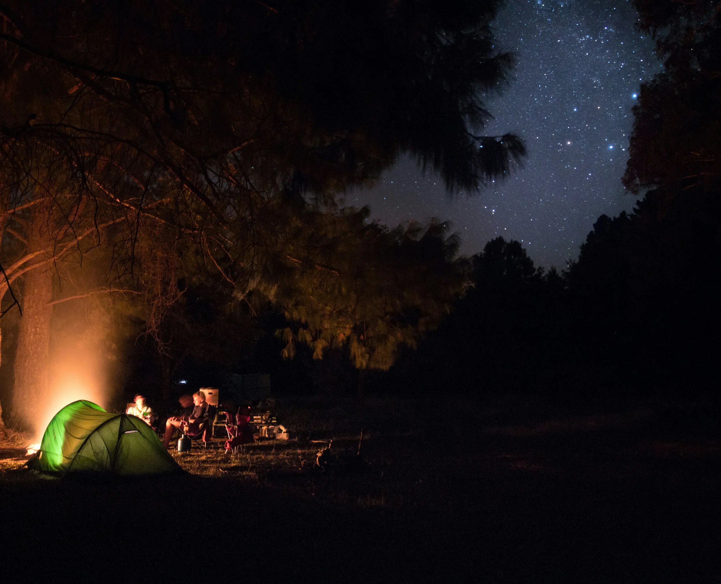 A group of travelers sitting around a glowing campfire in the dark Australian outback, with a crystal clear night sky full of stars and the Milky Way overhead.