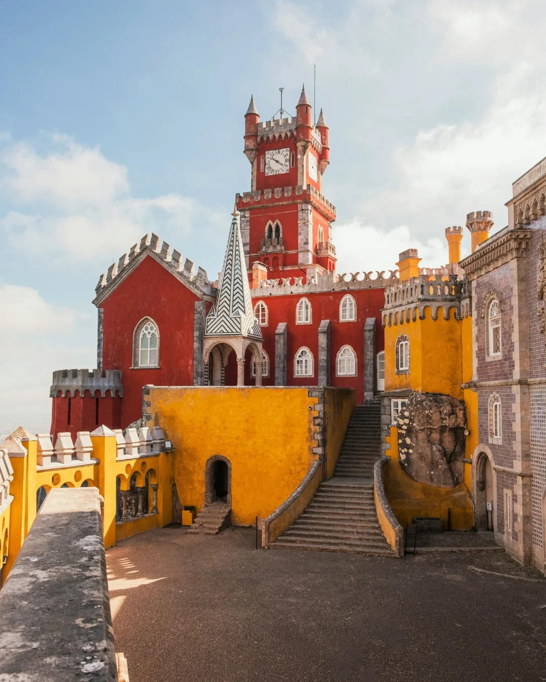 Colorful castle with red, yellow, and gray walls, featuring a tall clock tower with turrets and pointed roofs, under a partly cloudy sky.