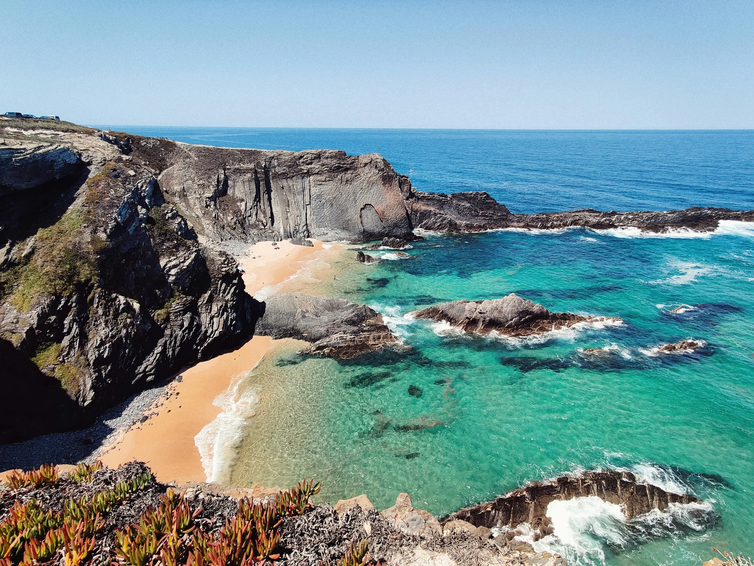 Scenic view of a wild sandy beach in Alentejo, Portugal, surrounded by high dark cliffs and the crashing waves of the blue Atlantic Ocean.