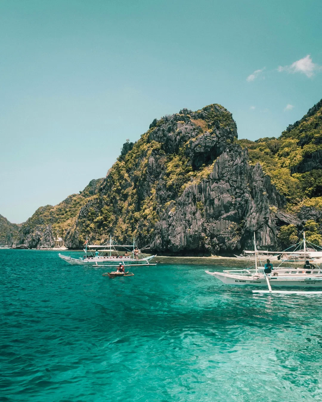 A scenic view of El Nido, Palawan in the Philippines, featuring turquoise waters, traditional white bangka outrigger boats, and towering limestone karst cliffs.