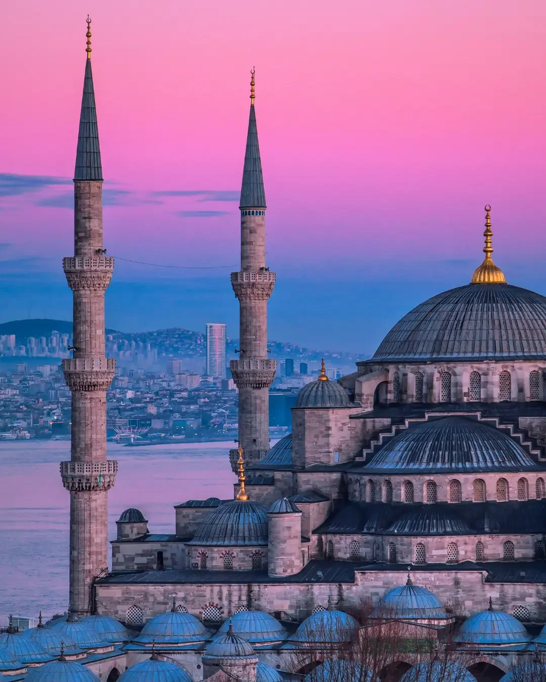 A mosque with multiple domes and two tall minarets against a sunset sky in a cityscape.