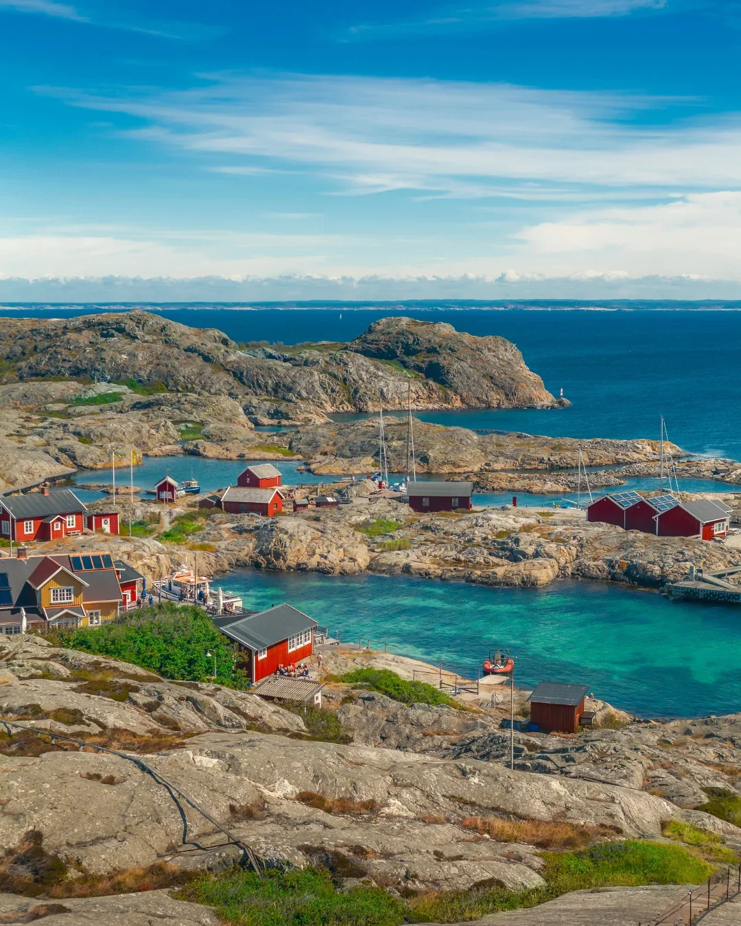 A high-angle landscape of the Väderöarna (Weather Islands) in Sweden, featuring traditional red wooden houses nestled among rocky cliffs and the turquoise waters of the Bohuslän archipelago.