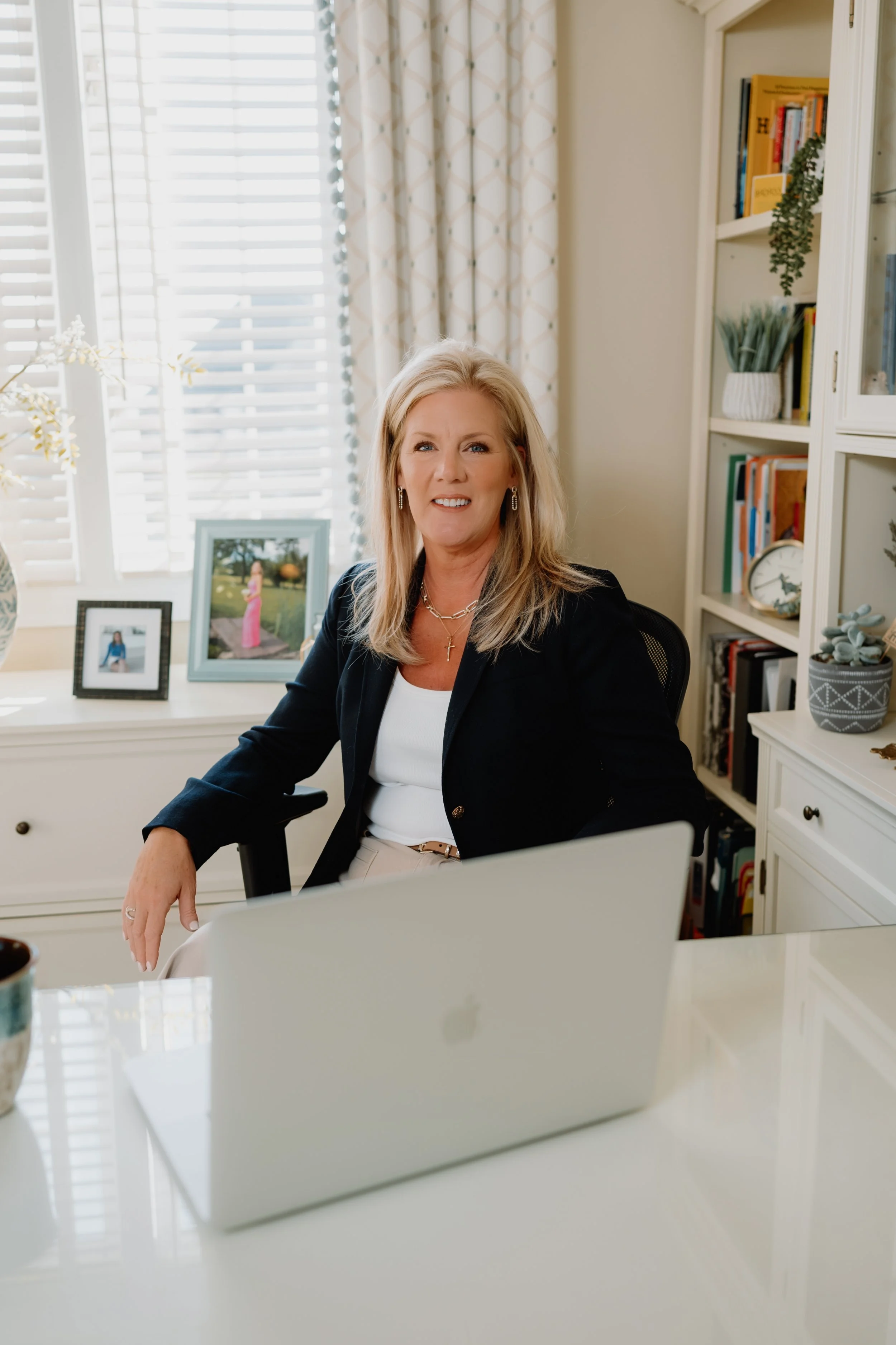 A woman with blonde hair, wearing a black blazer and white top, seated at a desk with a silver MacBook in front of her in a bright home office with window blinds, framed photos, and books.
