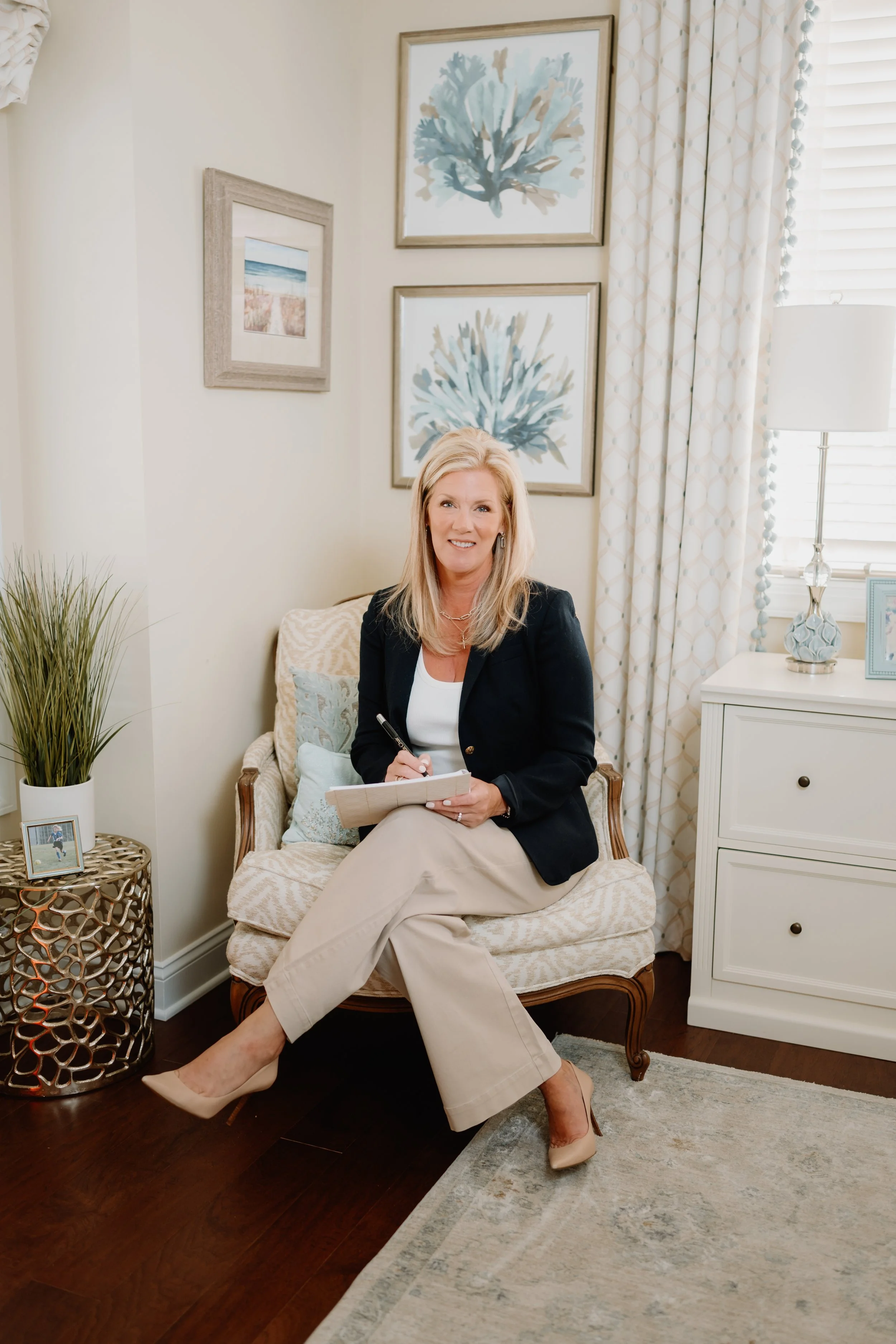 A woman with blonde hair sitting on a patterned beige armchair, holding a notepad and pen, in a well-decorated room with framed coastal artwork, a white nightstand, a lamp, a picture frame, and a tall green plant.
