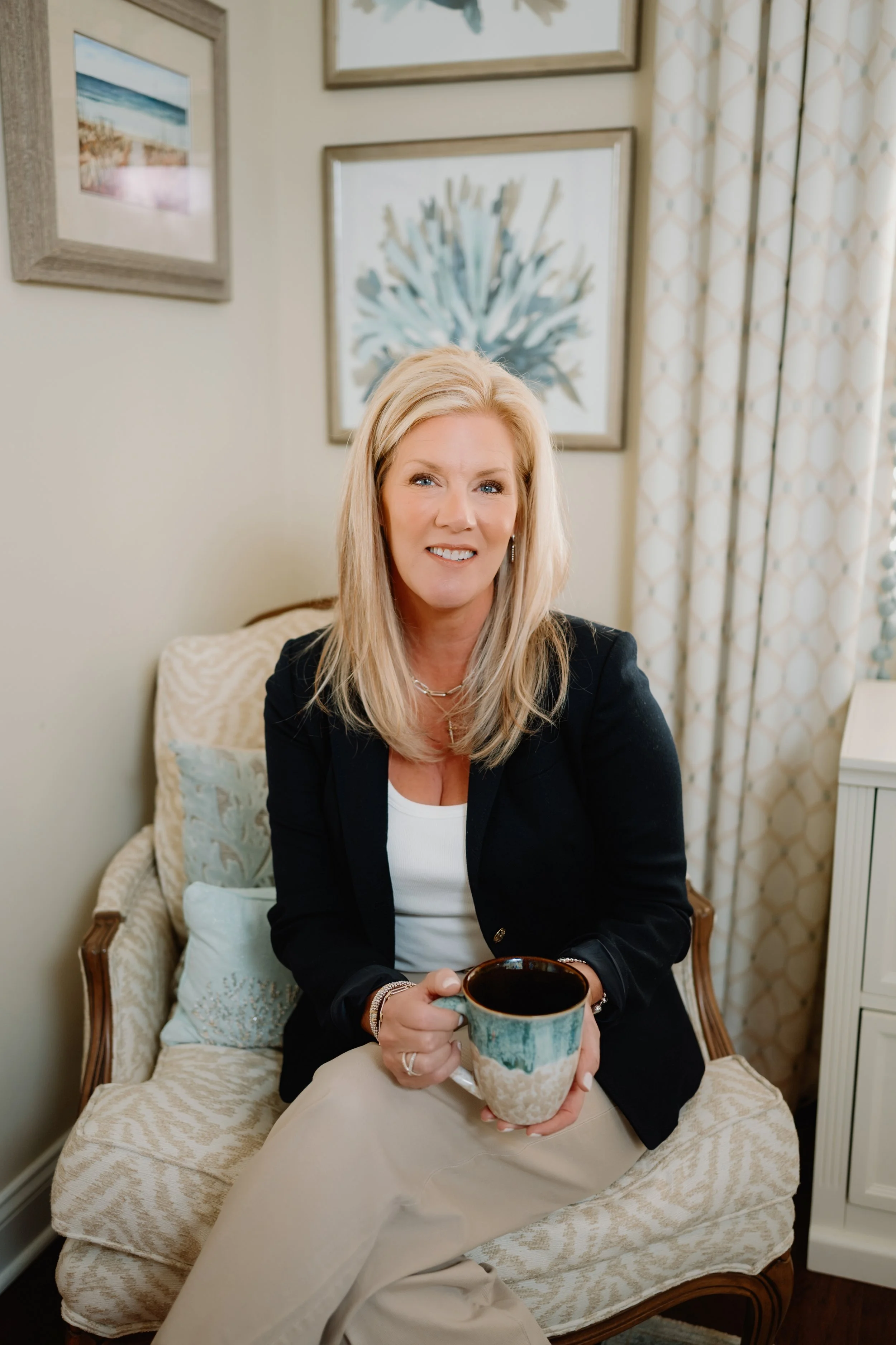 A woman with shoulder-length blonde hair, wearing a black blazer and white top, sitting on a beige patterned armchair, holding a ceramic mug, with framed artwork and curtains in the background.