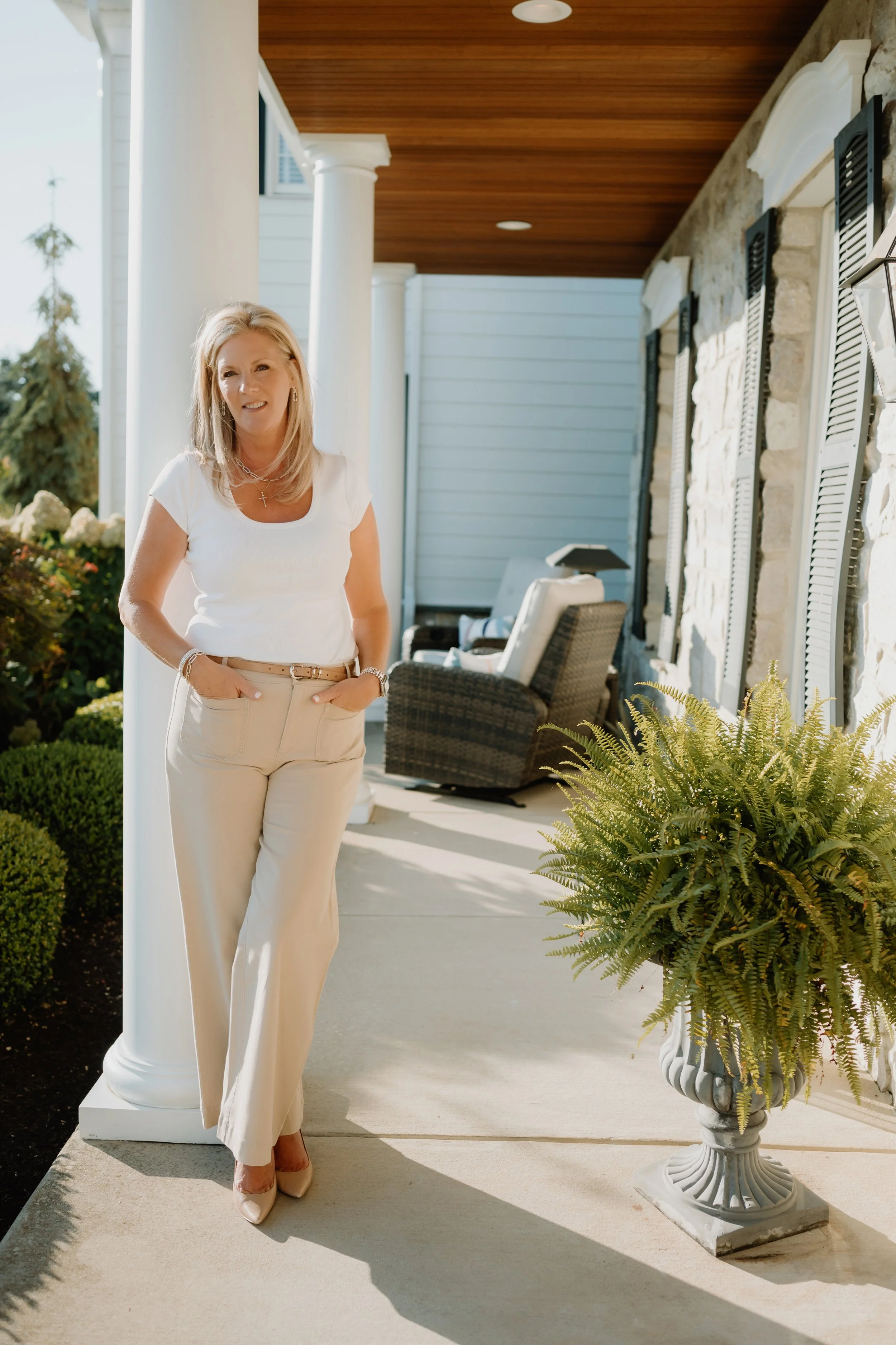 A woman with blonde hair, wearing a white t-shirt and beige pants, standing on a porch with a house and garden in the background.