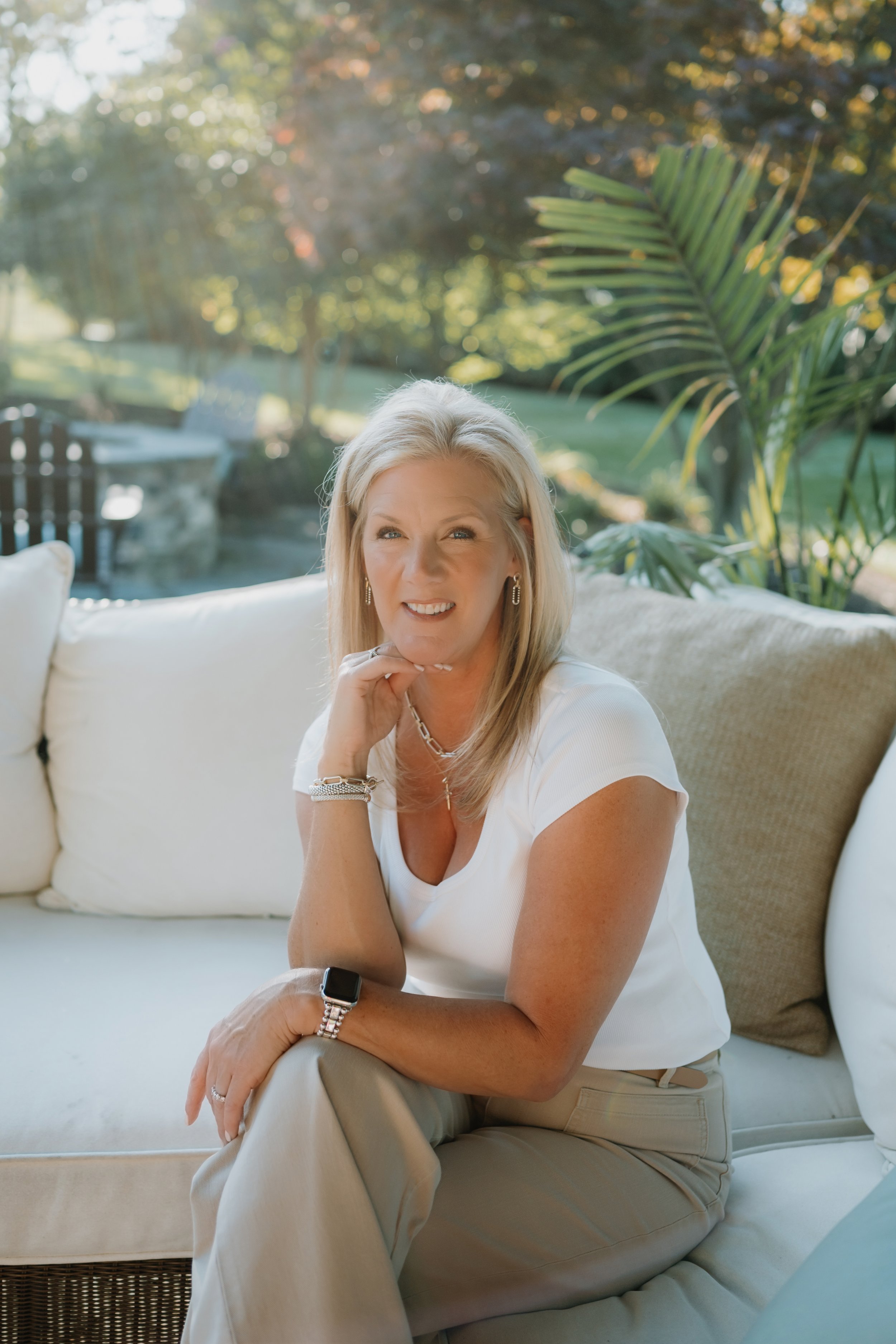 A woman with blonde hair sitting on a beige outdoor sofa, smiling at the camera, with a lush garden and sunlight in the background.