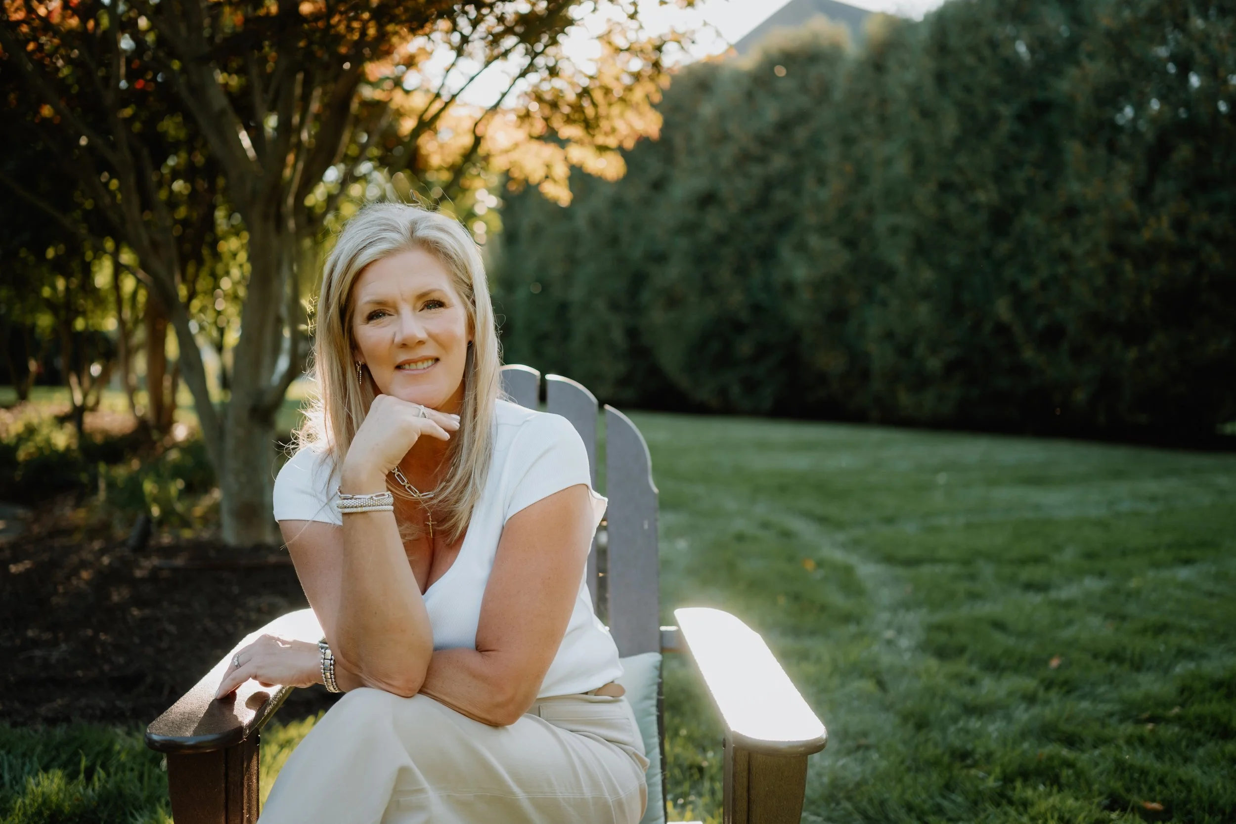 A woman with blonde hair sits on a bench in a park during sunset, wearing a white top and cream-colored pants, with trees and green grass in the background.