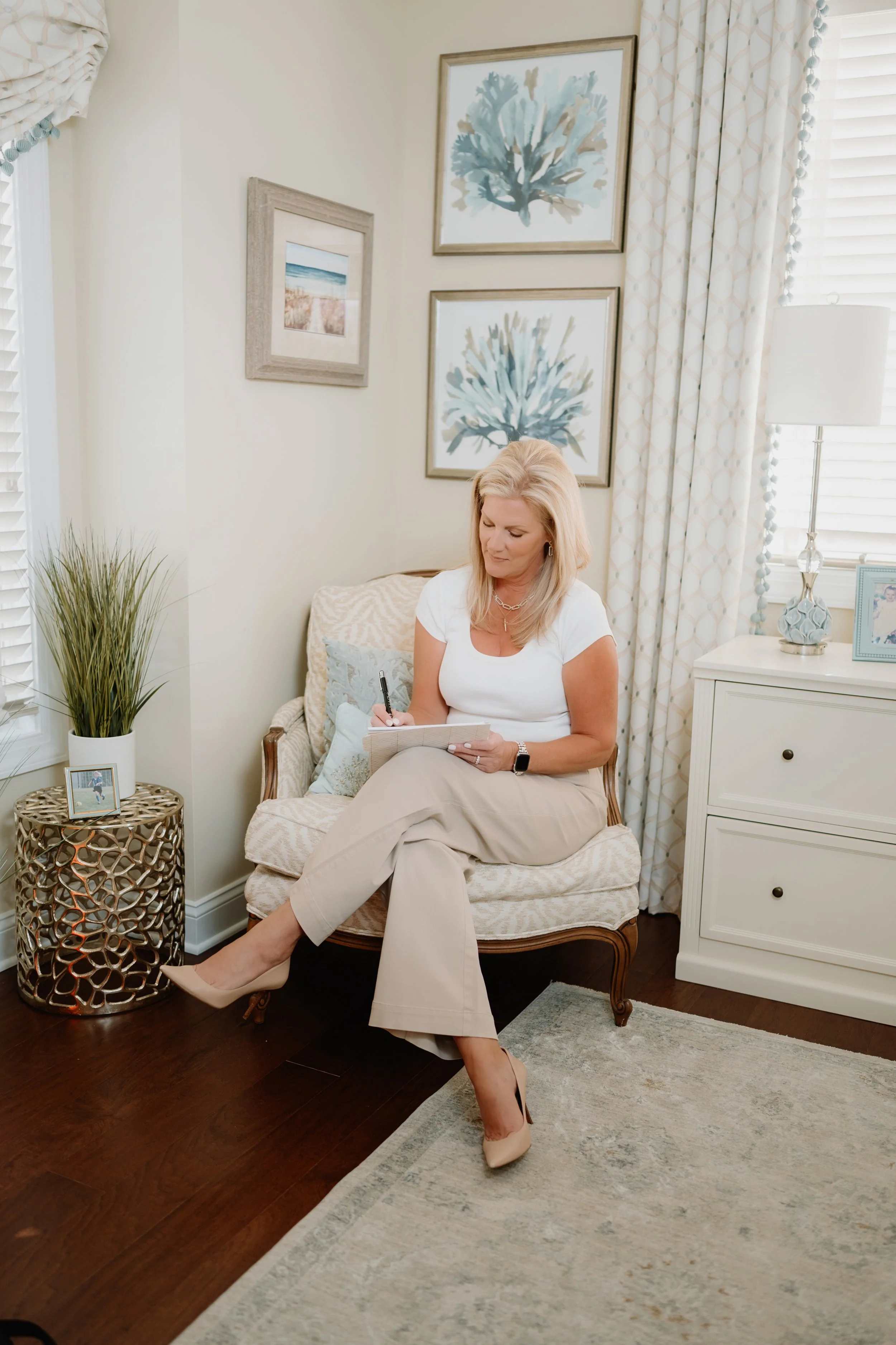 A woman with blonde hair wearing a white top and beige pants sitting on a cushioned armchair, writing on a notepad with a pen in a bright, cozy room decorated with framed wall art, a lamp, and a small table.