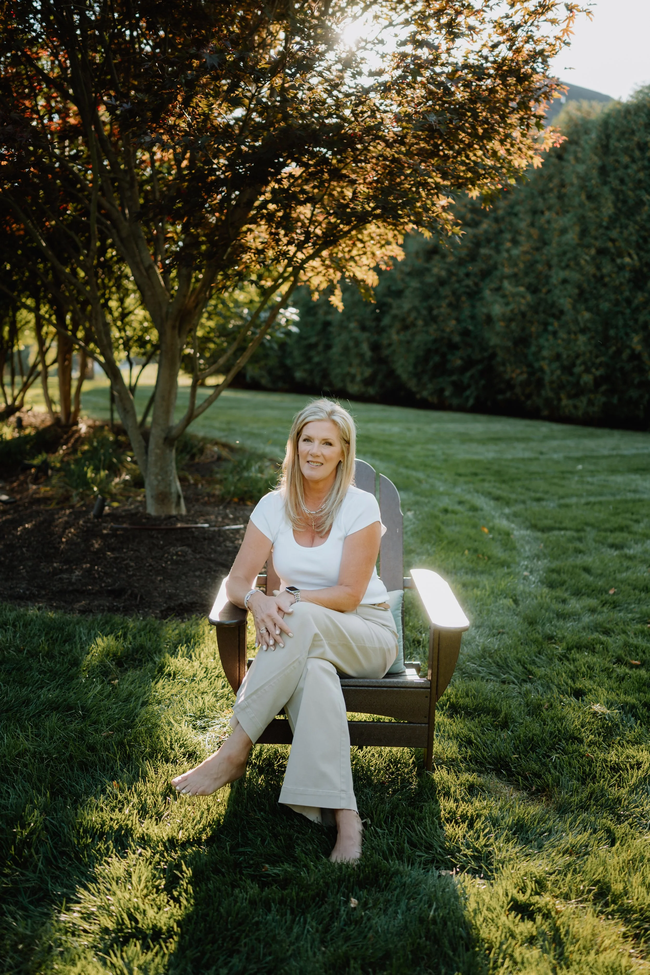A middle-aged woman with blonde hair sitting barefoot on a wooden outdoor chair in a garden, smiling, wearing a white shirt and beige pants, with trees and a grassy lawn in the background during late afternoon sunlight.