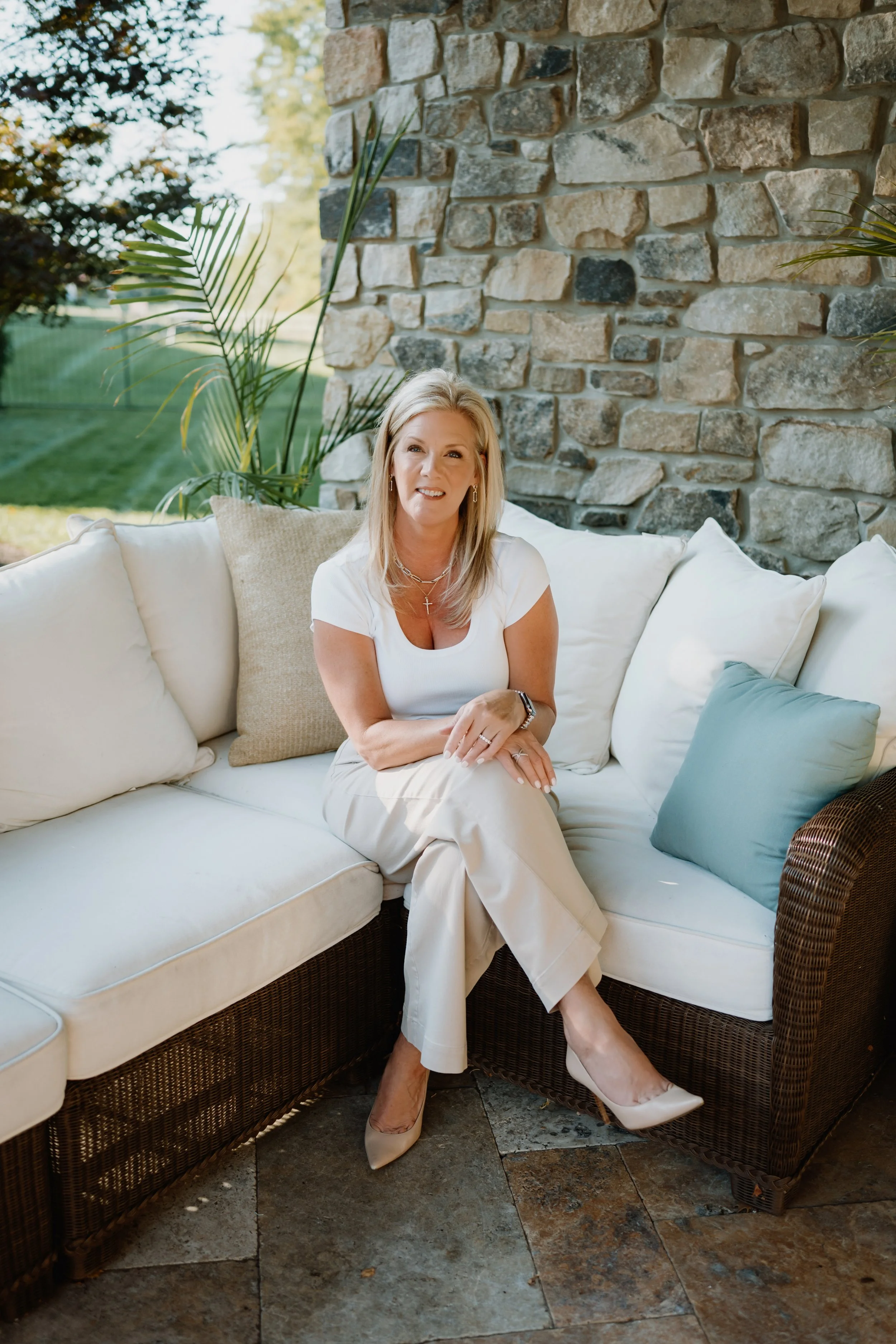 A woman with blonde hair sitting on a cream-colored outdoor sofa with pillows, in front of a stone wall, with green plants and a grassy yard in the background.