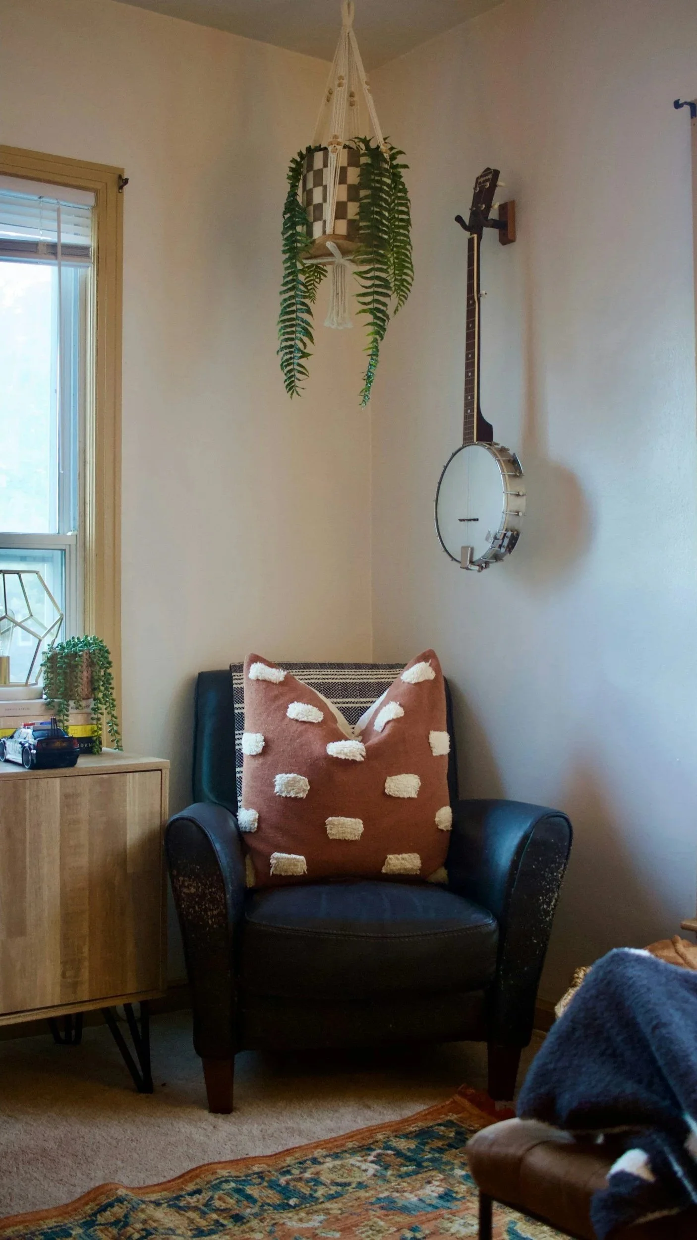 Cozy corner with a black armchair decorated with a pink pillow with white polka dots, a wooden side table with a small potted plant and toys, a window with blinds, and wall decorations including a hanging planter with lush green ferns and a banjo-shaped wall hanging.