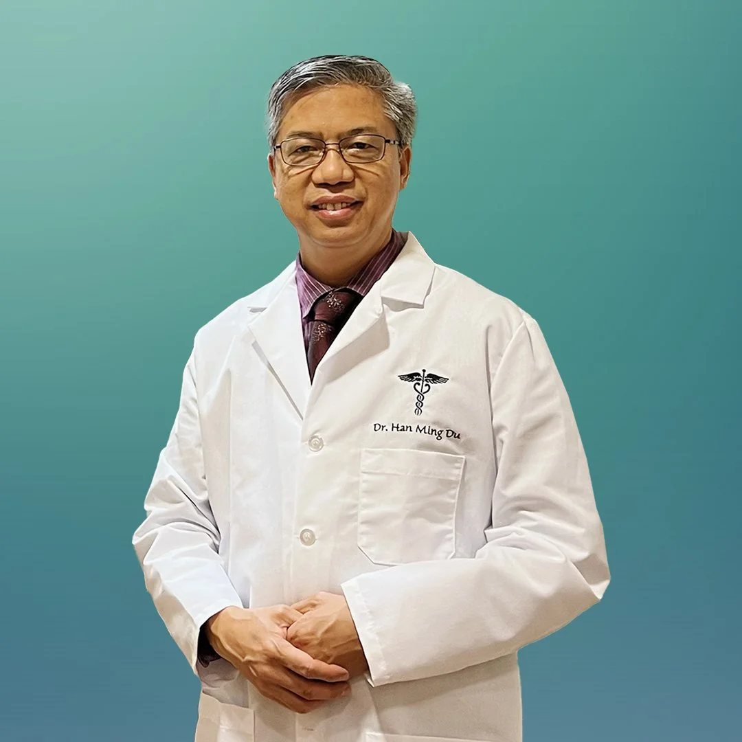 Dr. Han Ming Du, wearing glasses and a white doctor's coat, standing in his professional office.