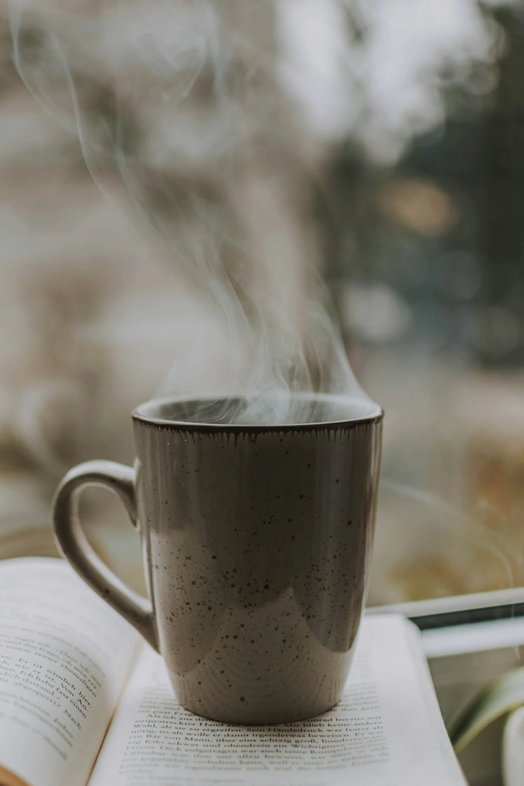 A steaming mug of coffee or tea on an open book, with a blurred window in the background.
