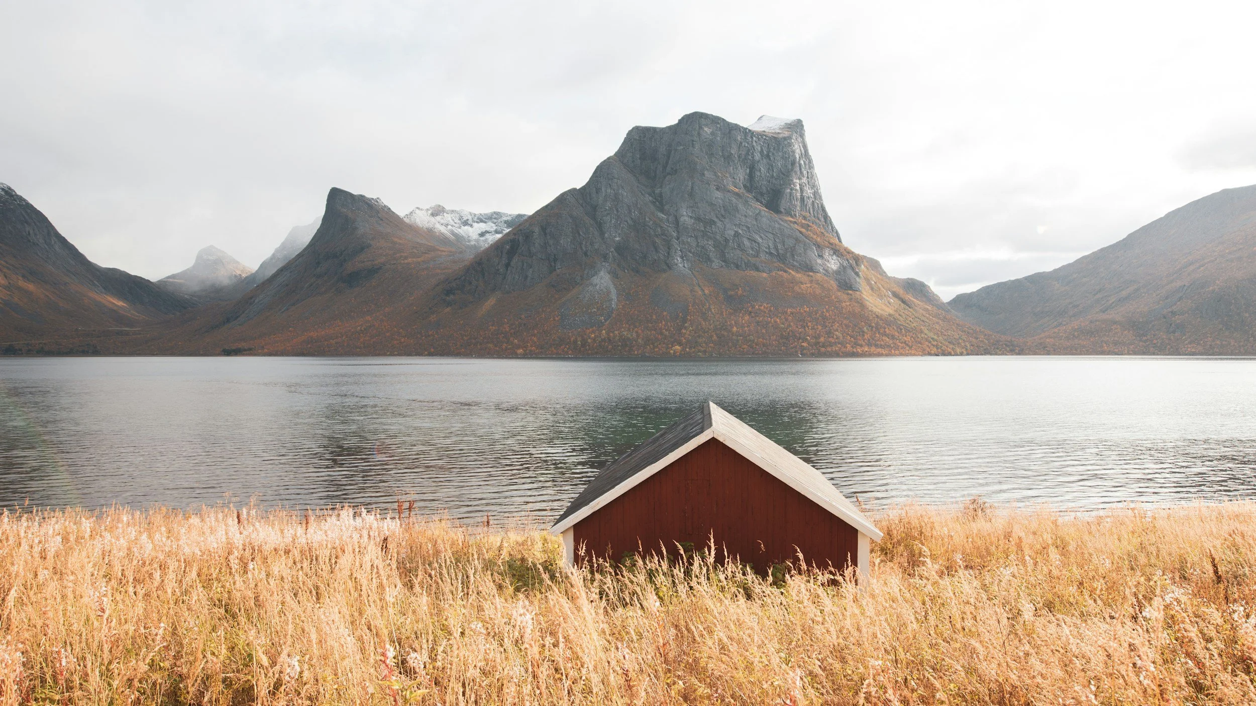 A small red and white boathouse on a grassy lakeshore with mountains in the background under a cloudy sky.