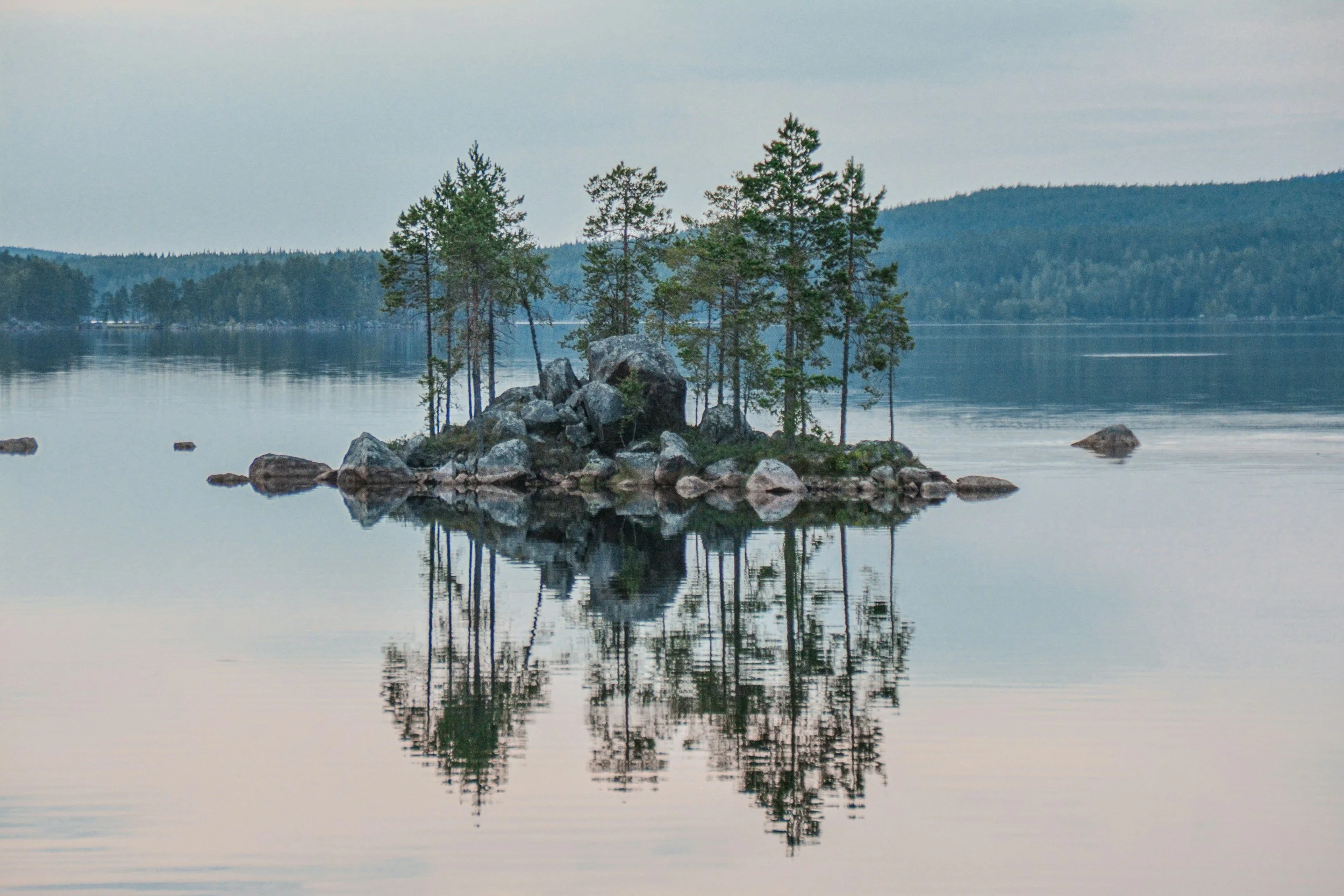 A small island with pine trees and rocks in the middle of a calm lake, reflecting the trees and rocks in the water, with distant hills in the background.