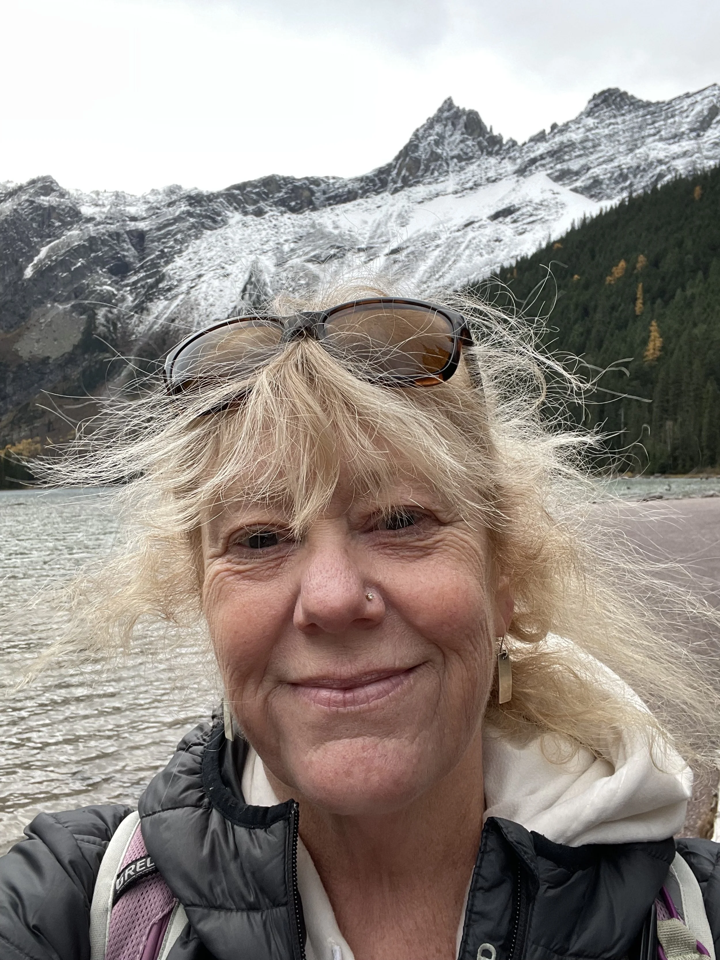 A woman with blond hair and sunglasses on her head taking a selfie outdoors near a river with snow-capped mountains and a forested hillside in the background.