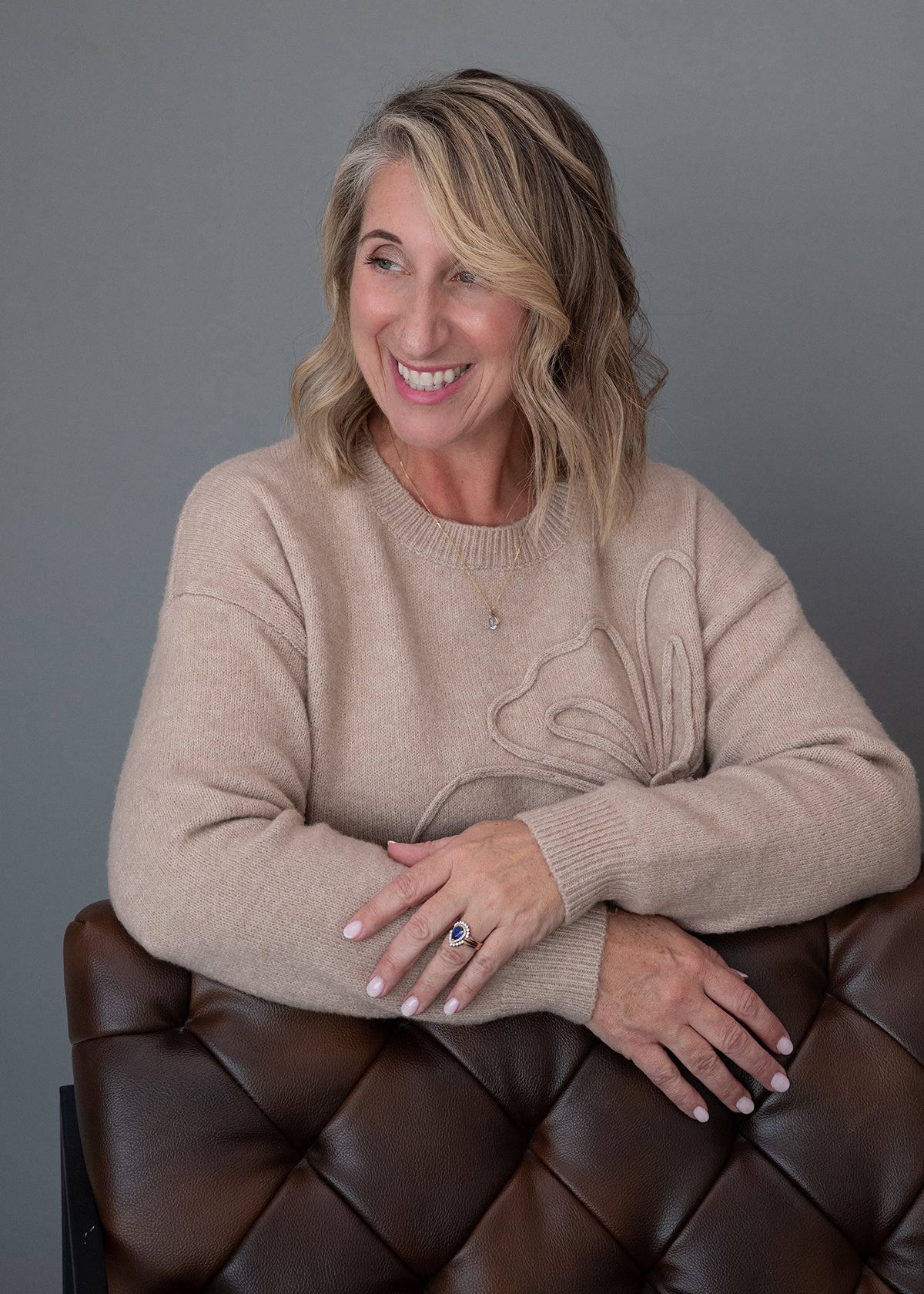 A woman with blonde wavy hair, smiling and looking to her left, sitting with her arms crossed on a brown leather chair, wearing a beige sweater with a floral pattern, a ring, and a necklace, against a plain gray background.