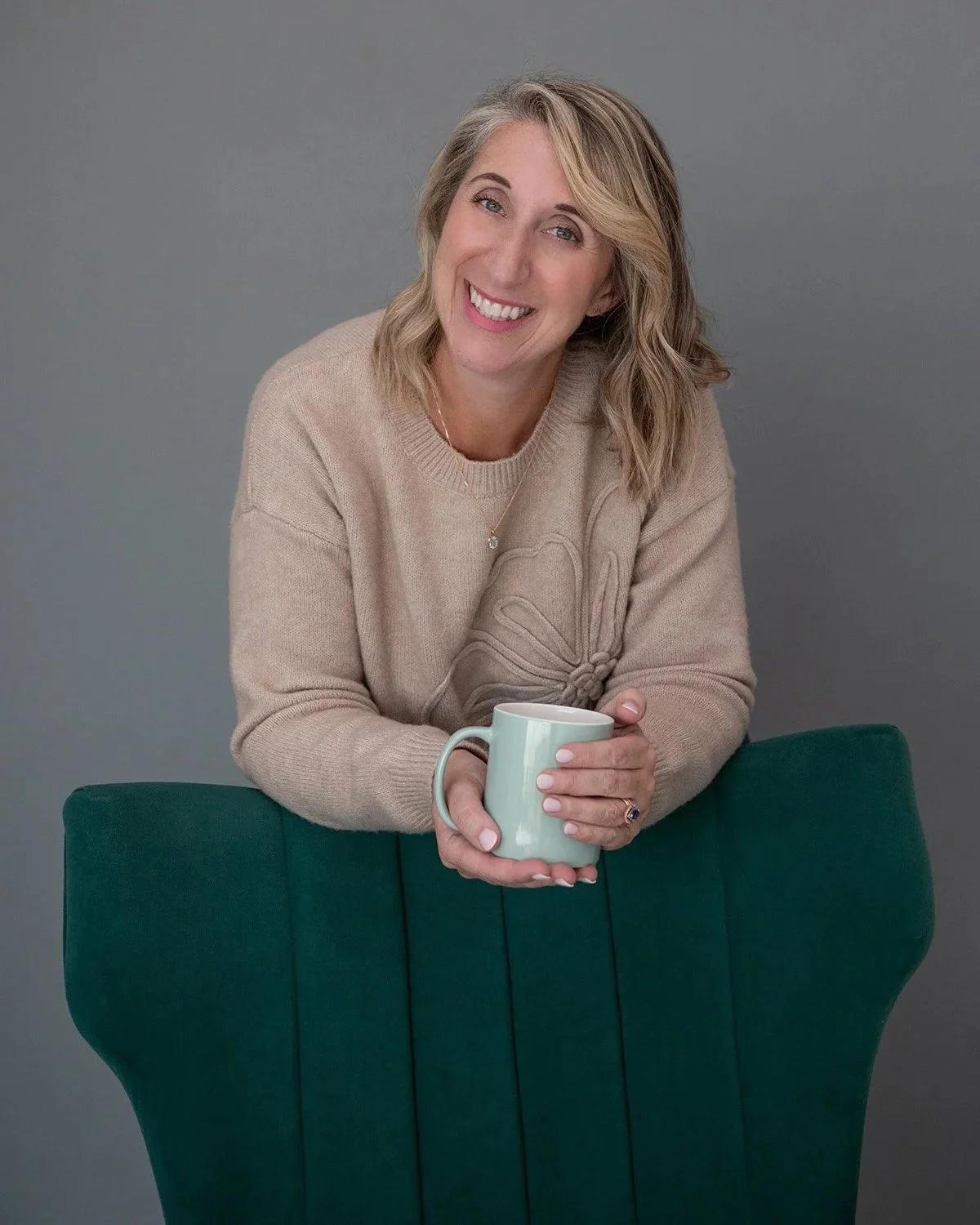 A woman with blonde hair smiling, holding a coffee mug, standing behind a green chair against a gray background.