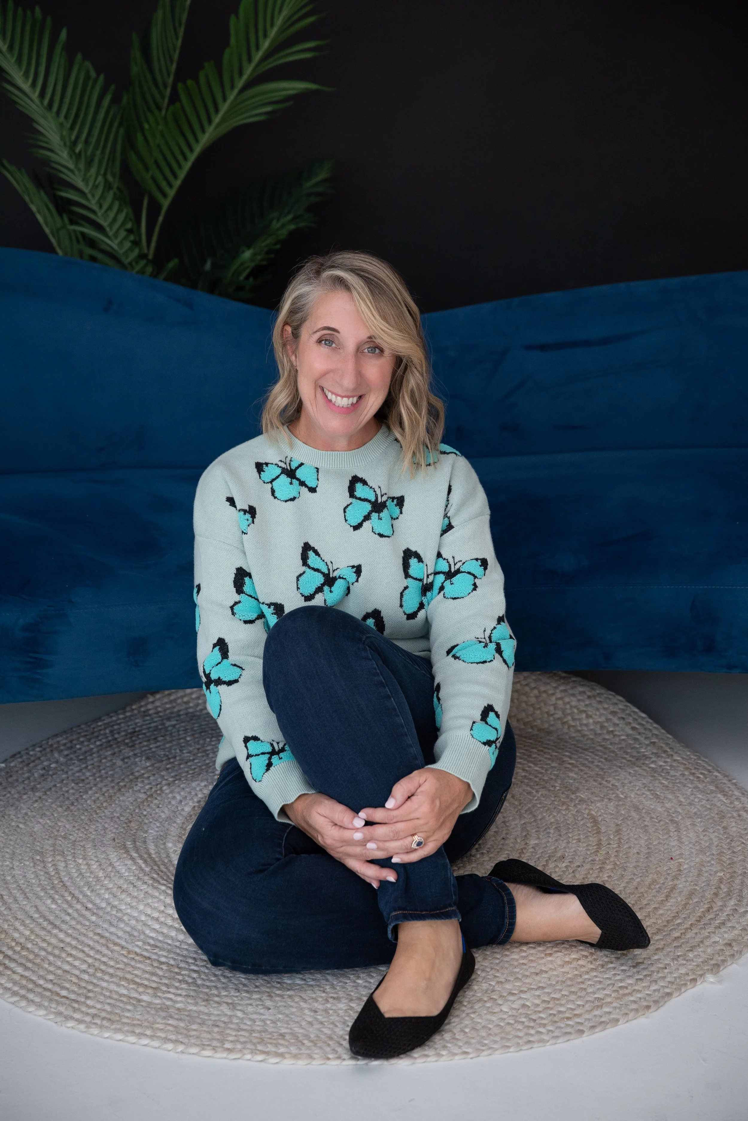A woman sitting cross-legged on a round beige rug, smiling, wearing a light green sweater with blue butterflies, dark jeans, and black flats, with a black wall, blue fabric, and green plant behind her.