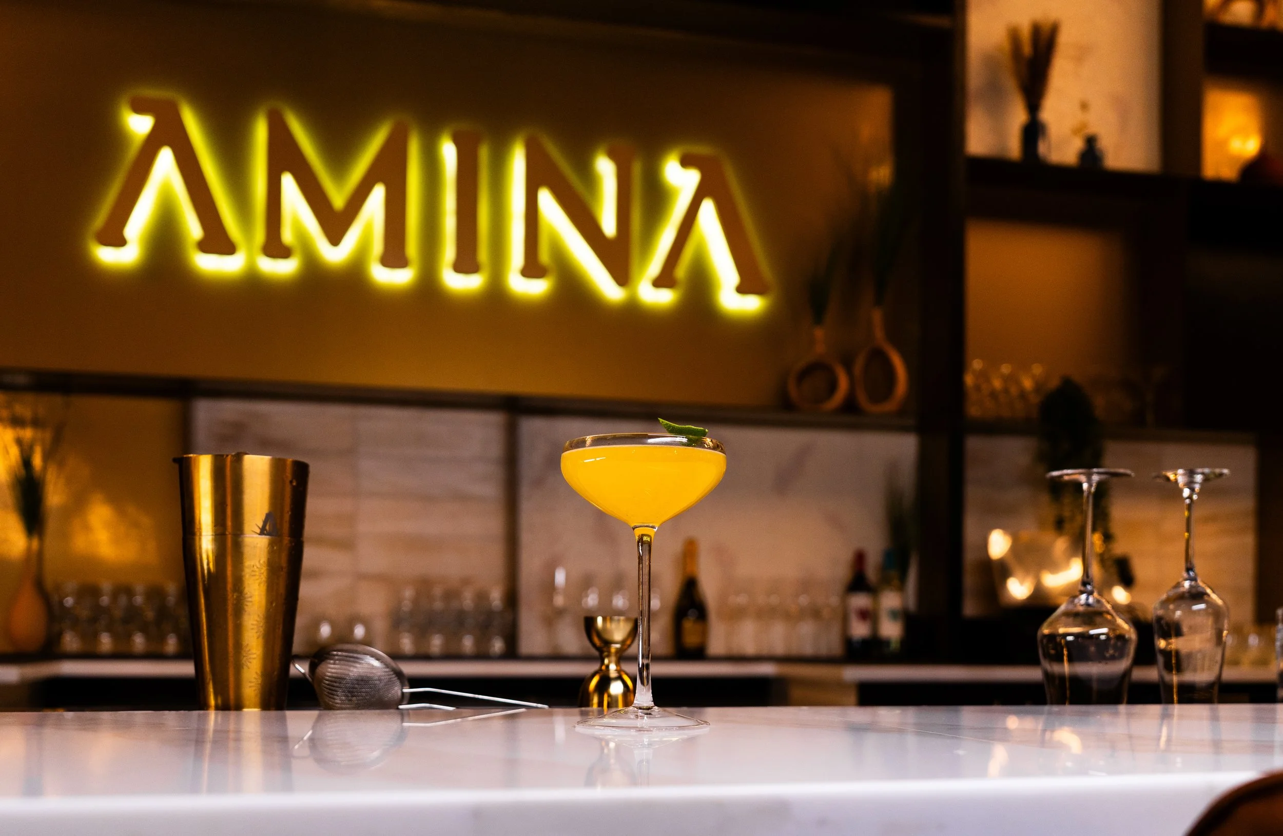A yellow cocktail drink with a garnish on a bar countertop with glasses and bar tools, with illuminated 'AMINA' sign in the background.