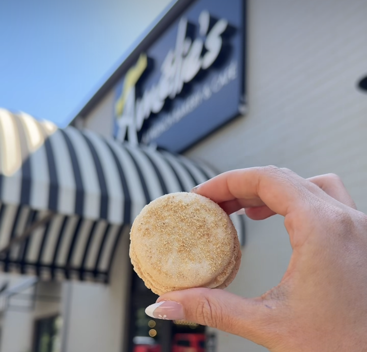 Person holding a round breaded snack, possibly a macaron or cookie, outside of a storefront with a striped awning.