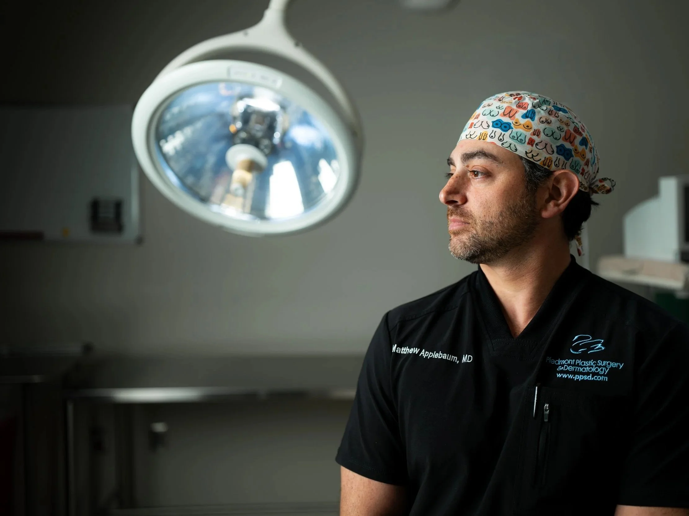 A doctor wearing surgical scrubs and a colorful cap sitting in an operating room, with a surgical light overhead.