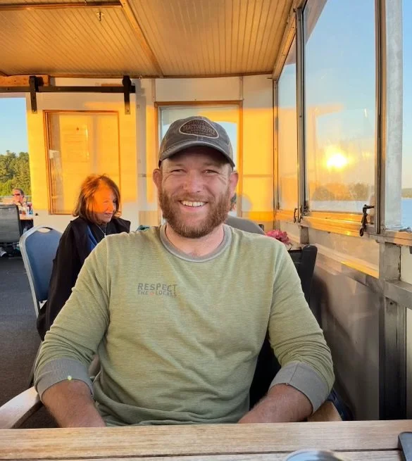 A man wearing a baseball cap and a light green shirt, smiling and sitting at a table in a restaurant or cafe during sunset, with a woman and a sunset view visible through windows in the background.