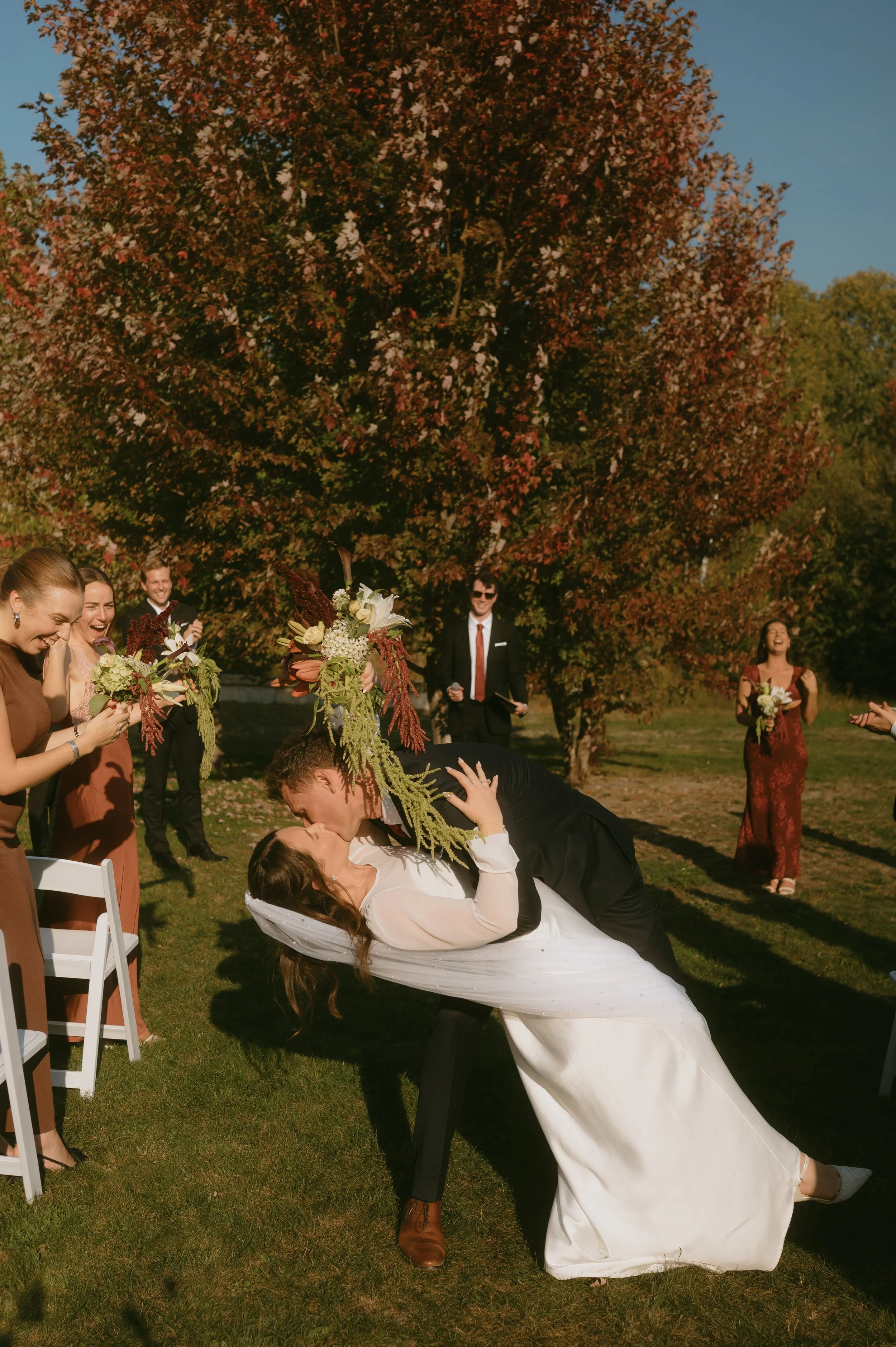 A bride and groom share a kiss outside during their wedding ceremony, with the groom dipping the bride. Guests watch and smile, some holding bouquets, under a large autumn tree.