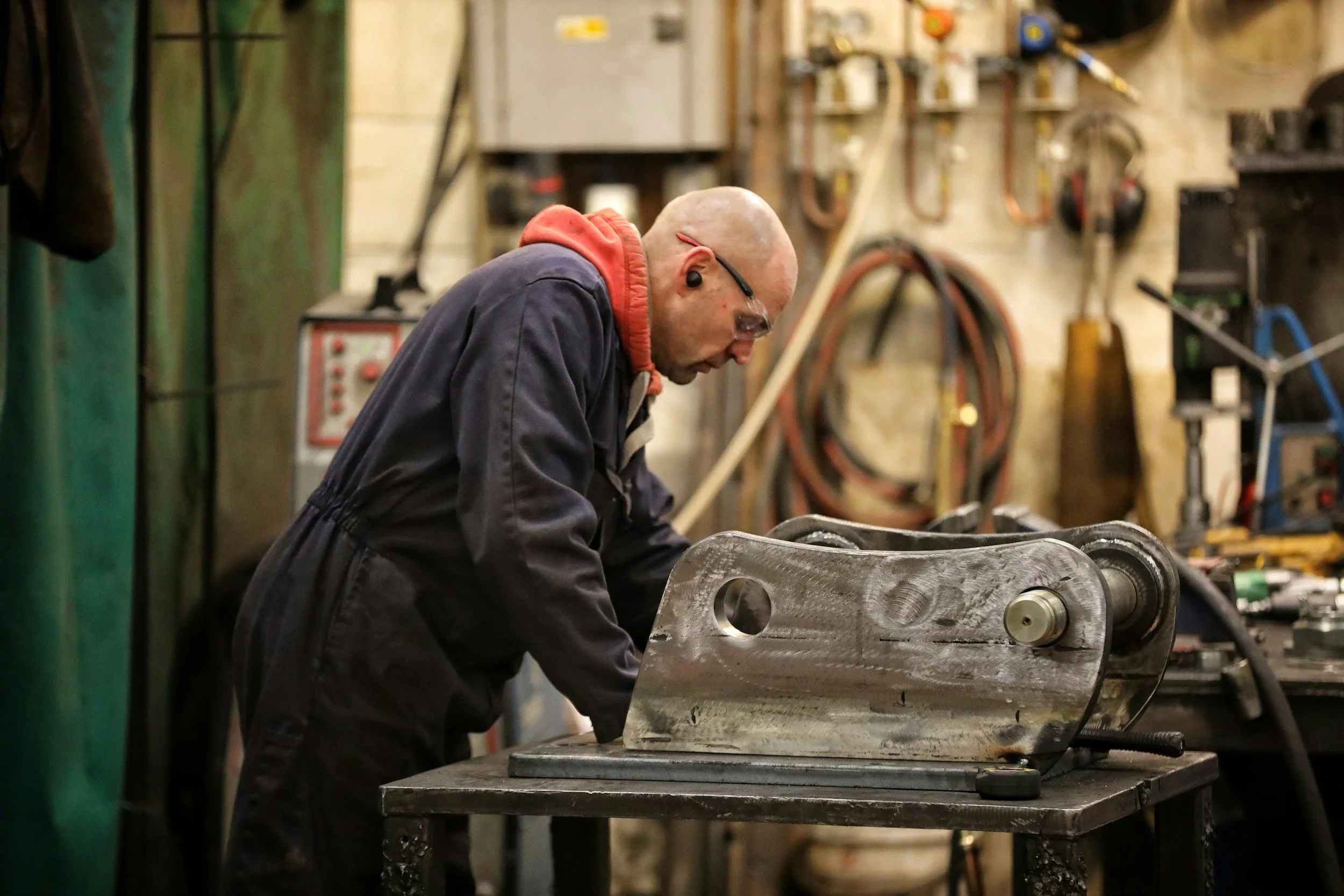 A man working on a metal object in a workshop, wearing safety glasses and ear protection, with tools and equipment in the background. GarageShops.ca