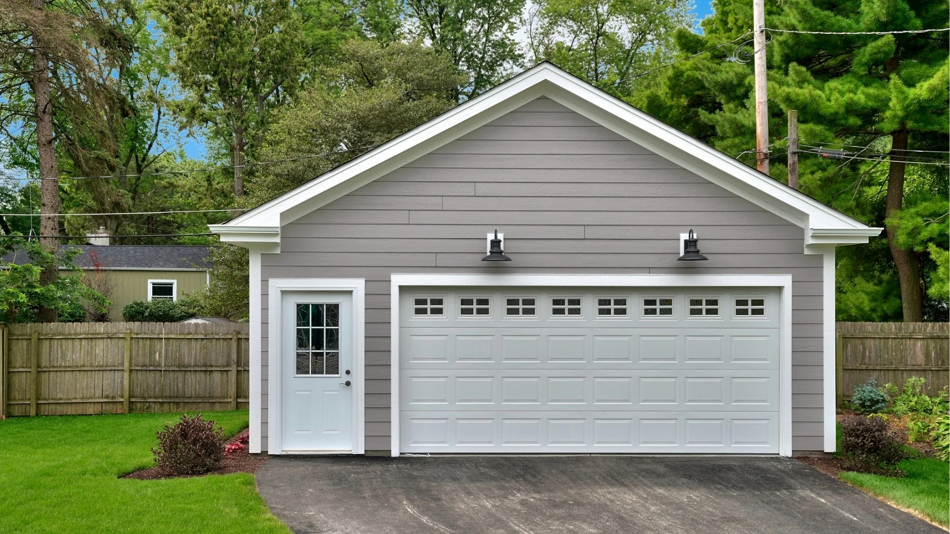 A gray garage with white door and small side door, black light fixtures, green lawn, wooden fence, and tall trees in the background.