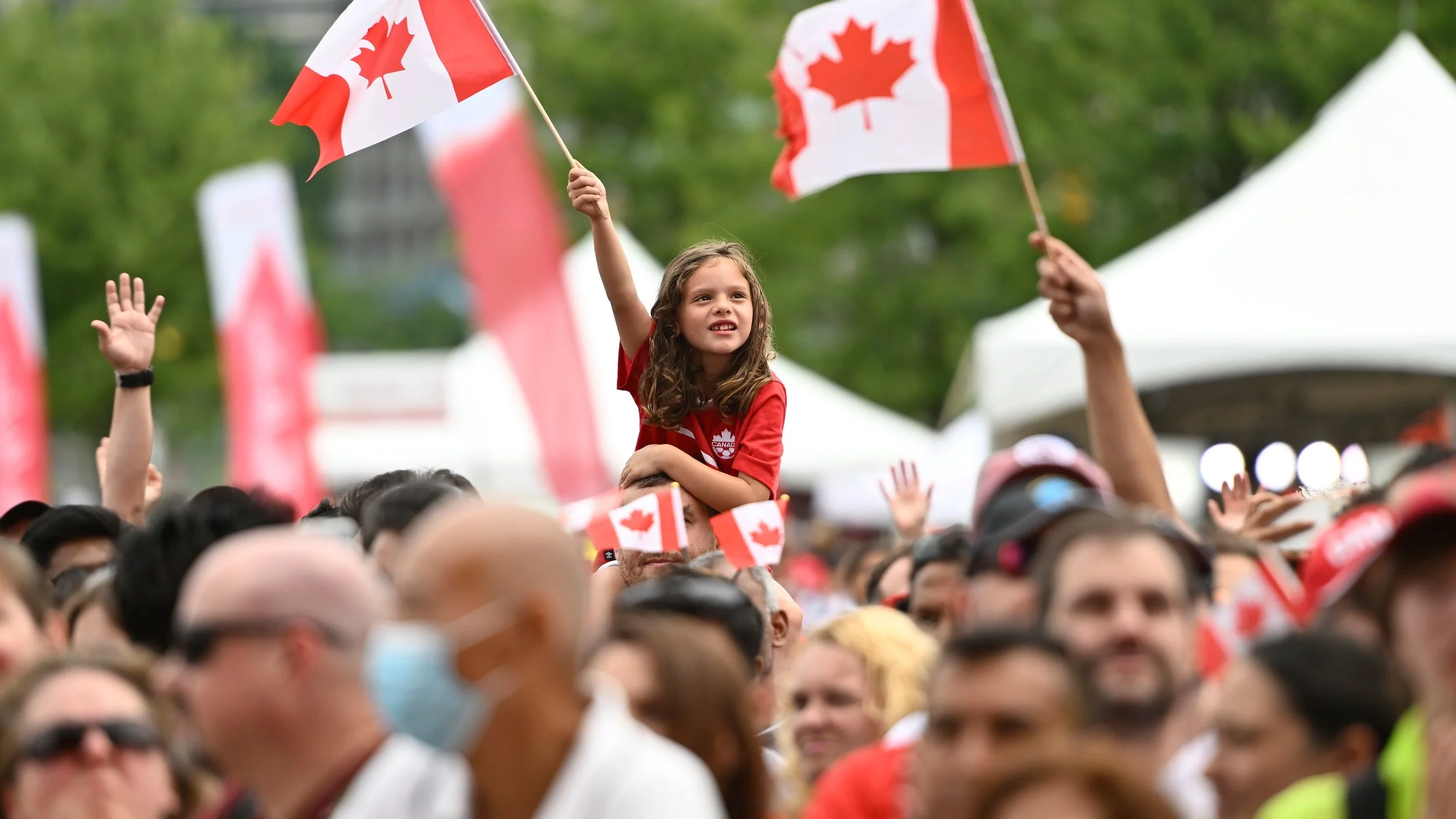 A young girl on someone's shoulders waving a Canadian flag at a crowd gathering, with many people waving flags and some wearing red and white clothing, likely at a patriotic or national celebration event.