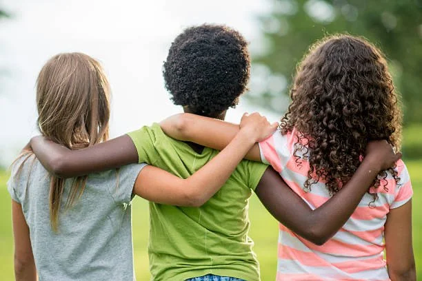 Three children with different skin tones, standing outside with their arms around each other, looking into the distance on a sunny day.