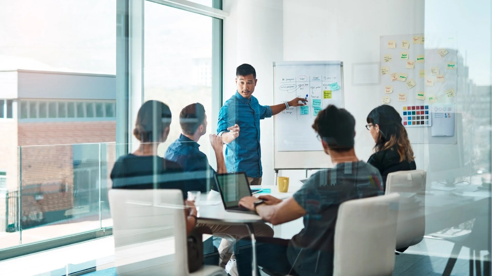 A man leading a business meeting or presentation with five colleagues seated around a table in a conference room. The man points to a diagram on a whiteboard, and the others are listening or taking notes with laptops and tablets. The room has large windows and sticky notes on the walls.