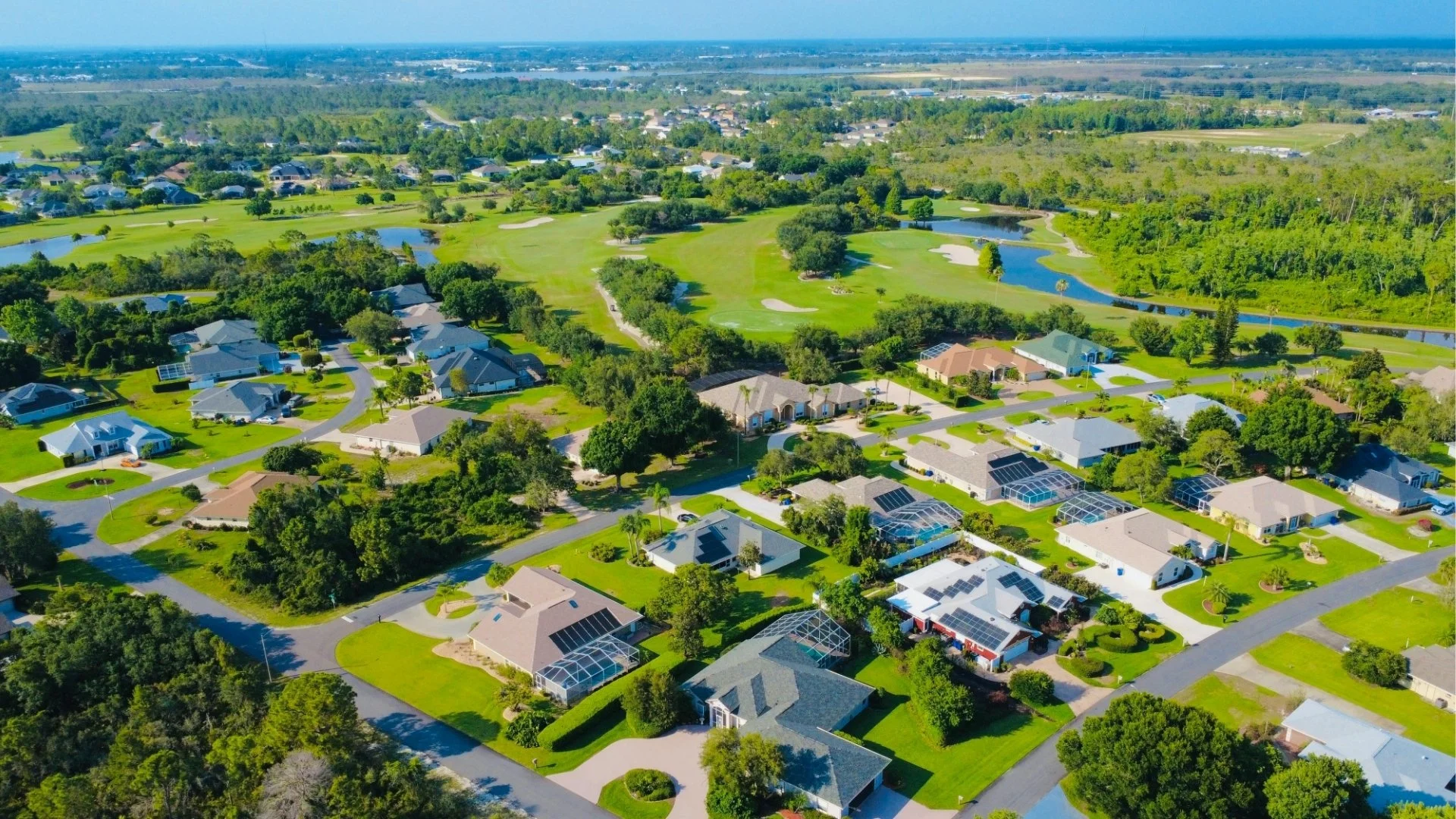 Aerial view of a suburban neighborhood with houses, green lawns, and a golf course with lakes in the background.
