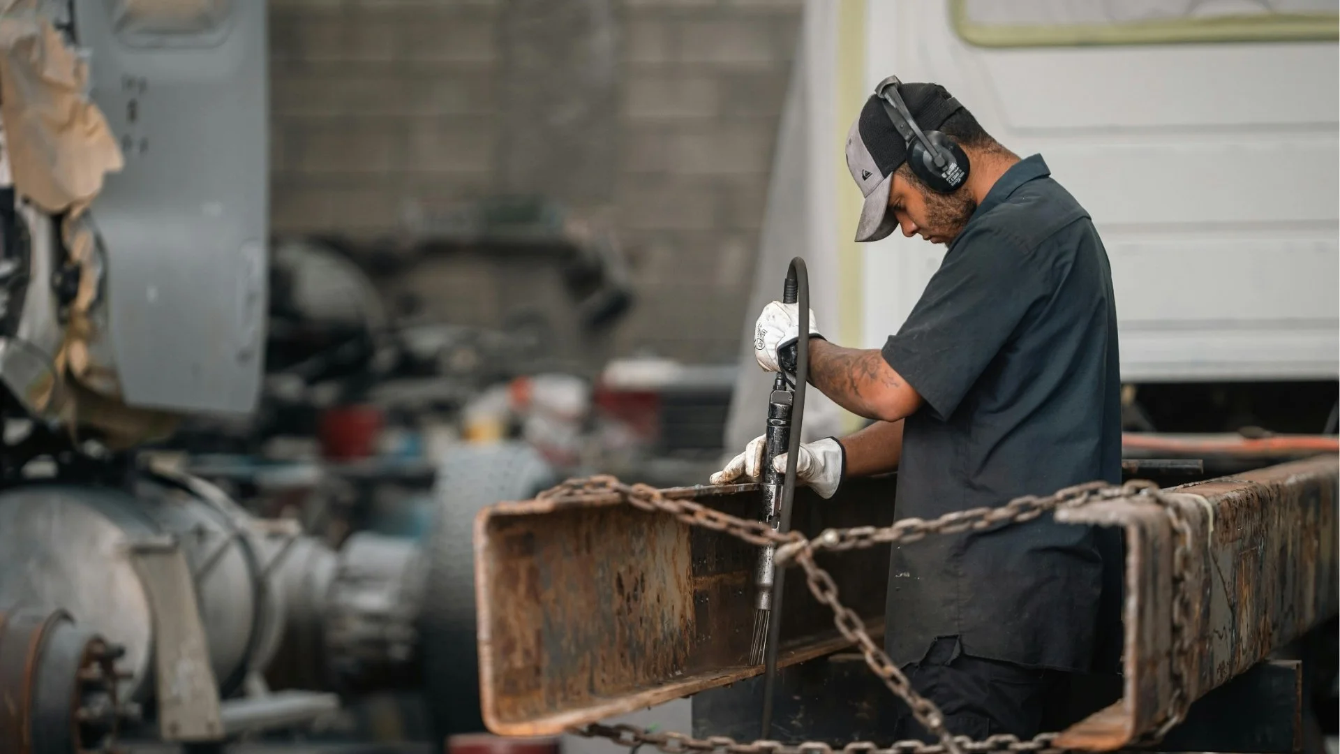 A man wearing a black shirt, gray cap, safety headphones, and gloves is working with a hydraulic tool on a rusty metal surface in an industrial workshop. GarageShops.ca