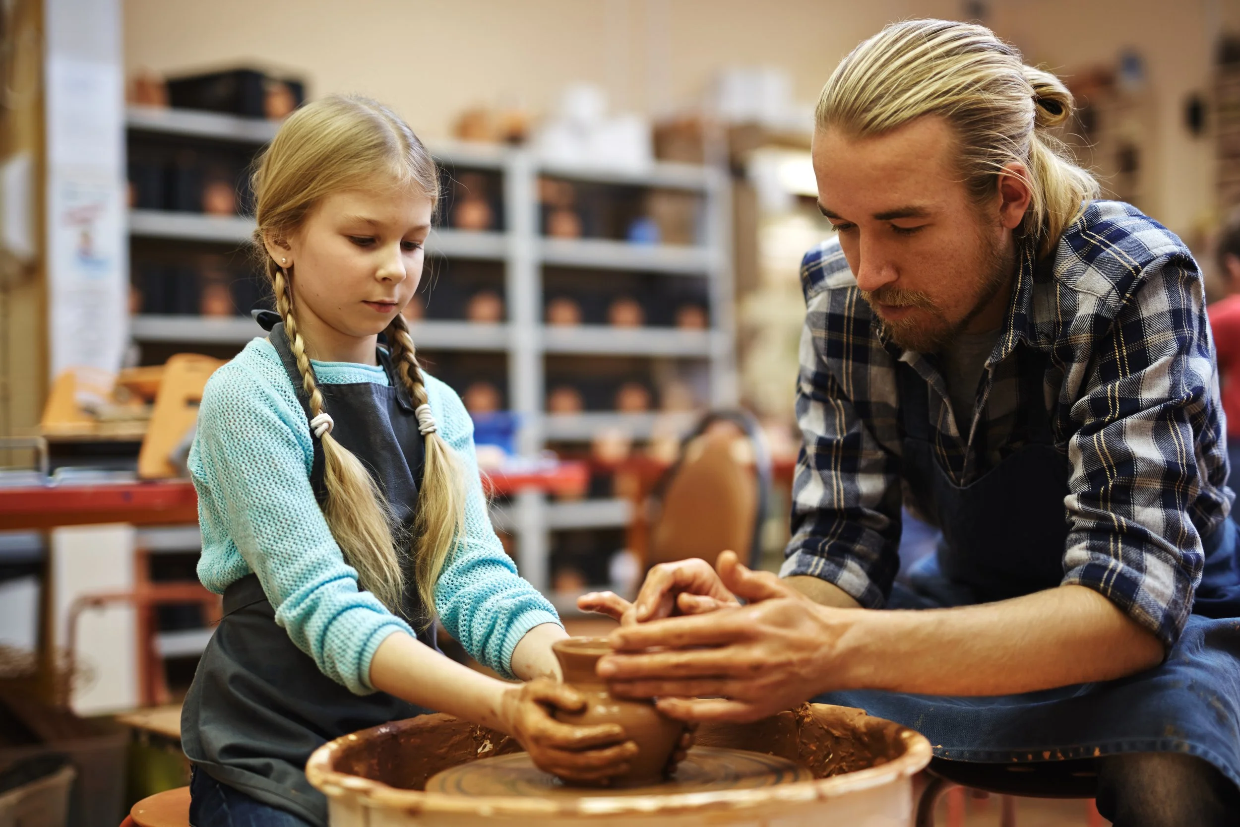 A young girl and a man working together on a pottery wheel, shaping clay into a vessel in a pottery studio.