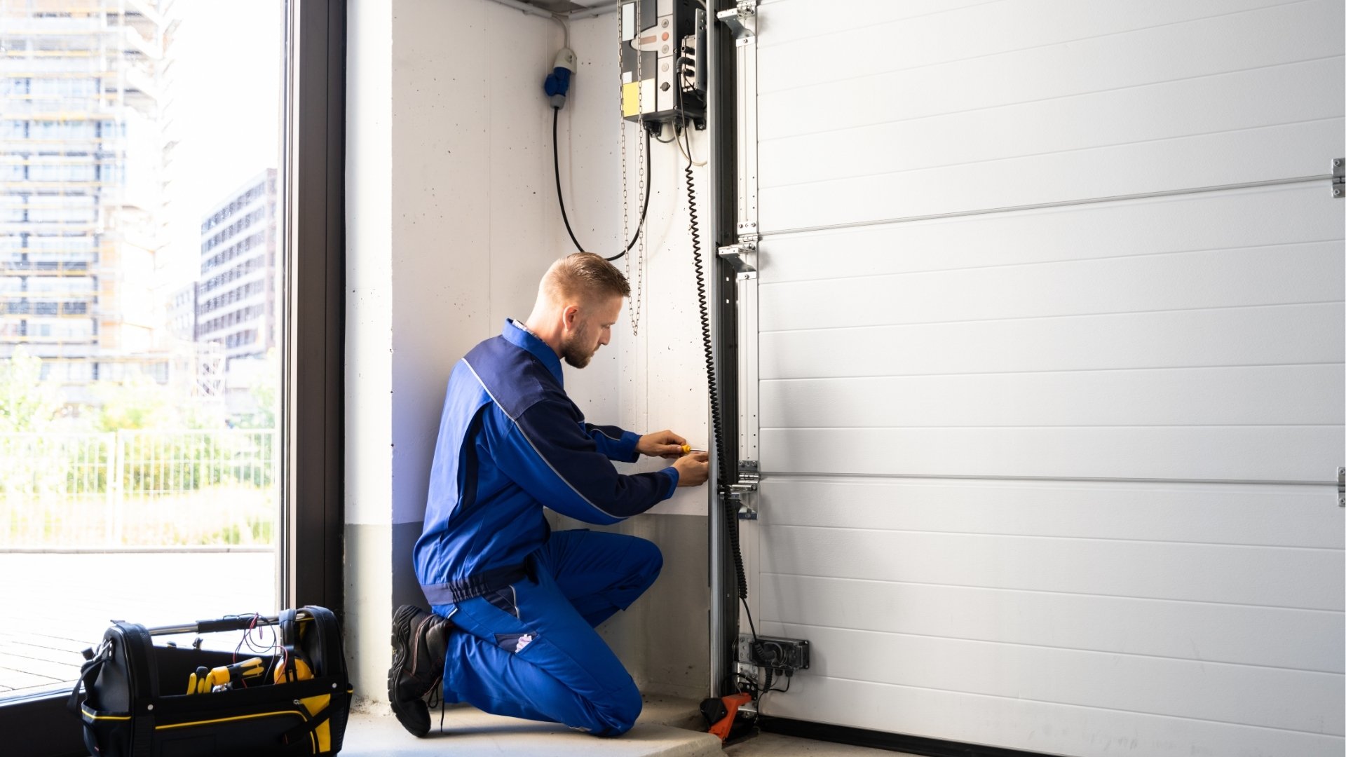 A technician kneels on the floor in blue work clothes, working on the mechanism of a large industrial GarageShops.ca optically aligned with tools in a black and yellow tool bag beside him, in a bright room with city buildings visible outside window.