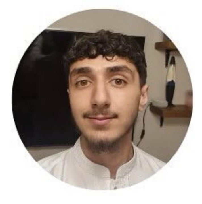 A young man with curly dark hair, light skin, and a slight mustache, wearing a white button-up shirt, smiling in a room with a shelf and mounted TV in the background.