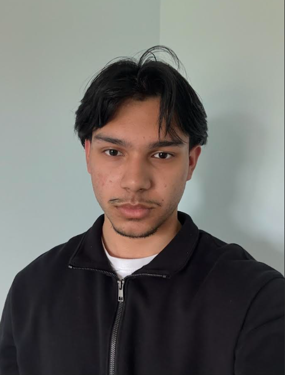A young man with dark hair, light brown skin, and facial hair, wearing a black zip-up jacket and a white shirt underneath, standing against a light-colored wall.