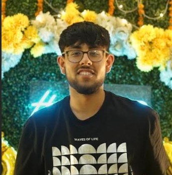 Young man with glasses smiling at a decorated event with yellow and white flowers and green foliage in the background.