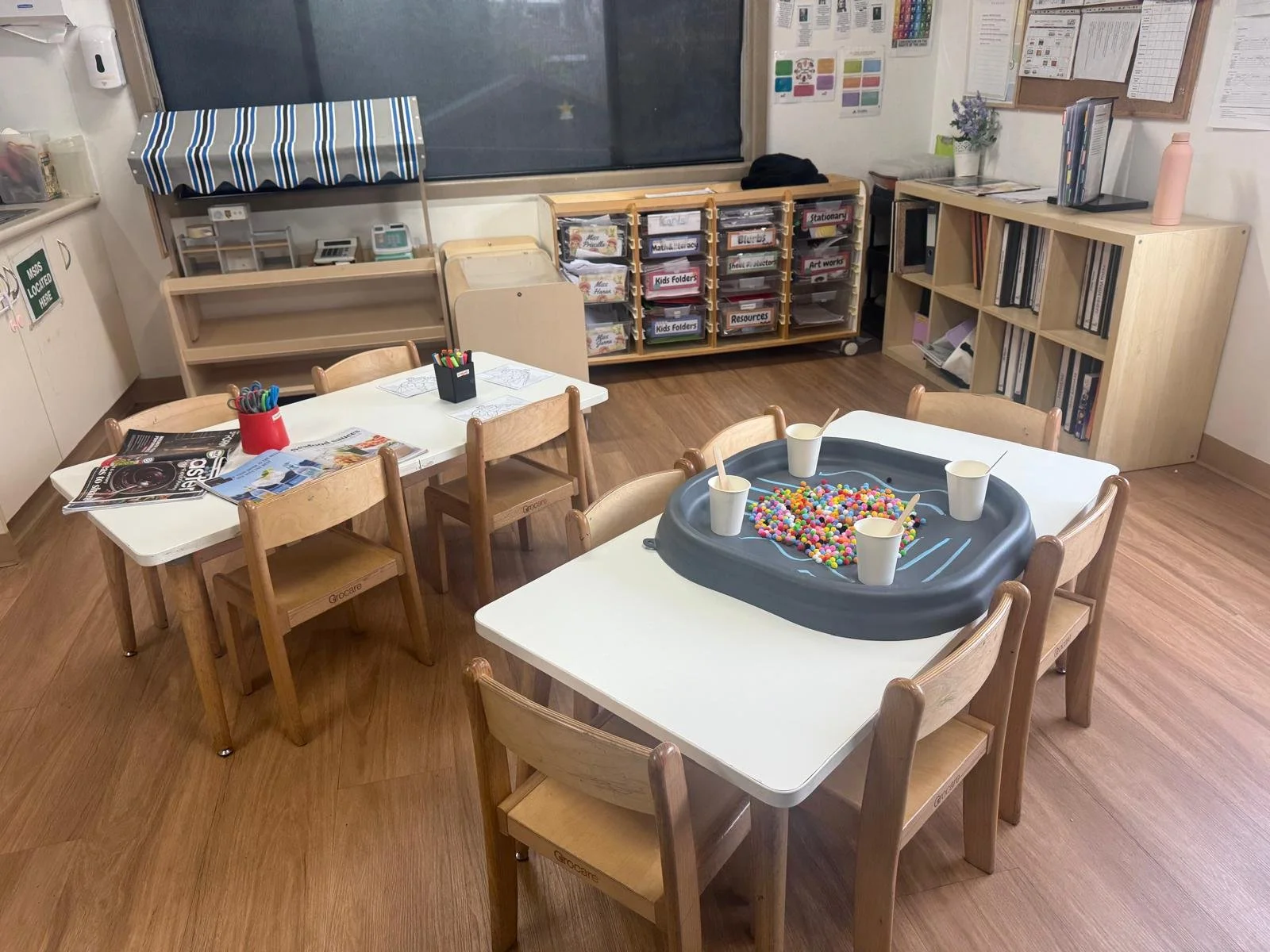 An indoor classroom with two white tables and multiple wooden chairs. One table has a black tray with cups, paintbrushes, and colorful beads; the other has a red container with markers, magazines, and papers. The room has wooden flooring, storage she